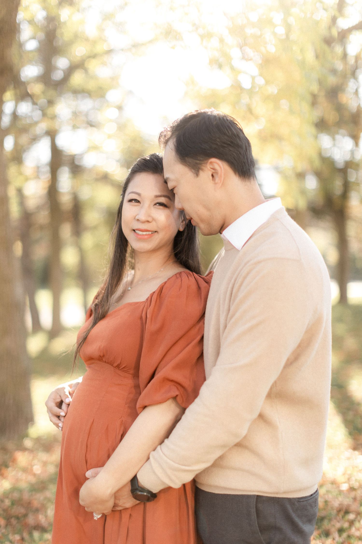 Intimate maternity photo of couple embracing in sunlit wooded park setting.