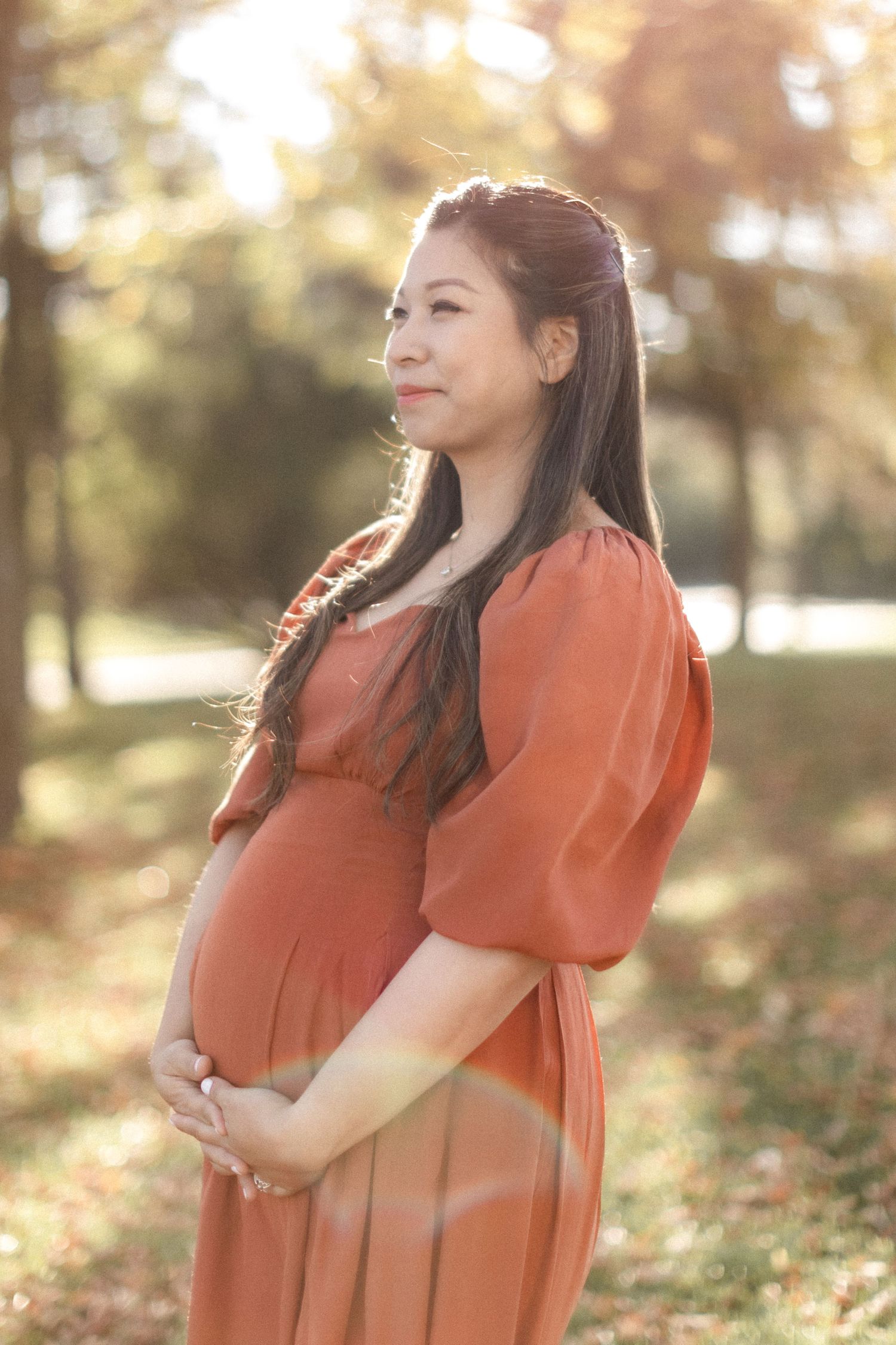 Side profile of an expectant mother in a rust-colored dress standing in dappled sunlight.