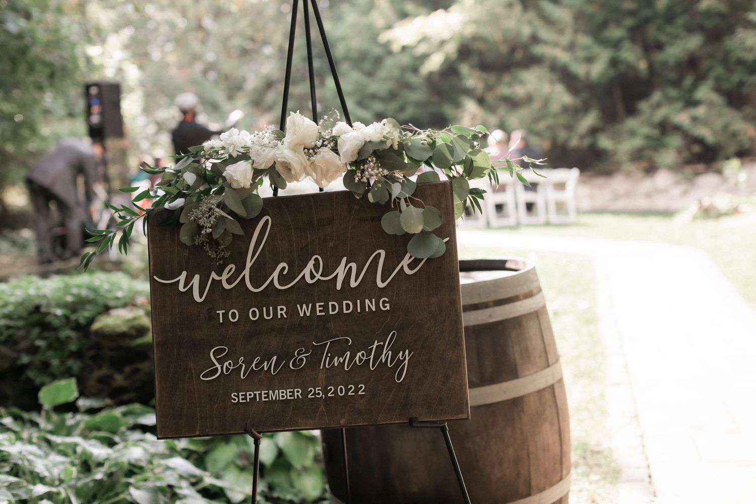 Rustic welcome sign on wooden barrel decorated with white flowers at outdoor wedding.
