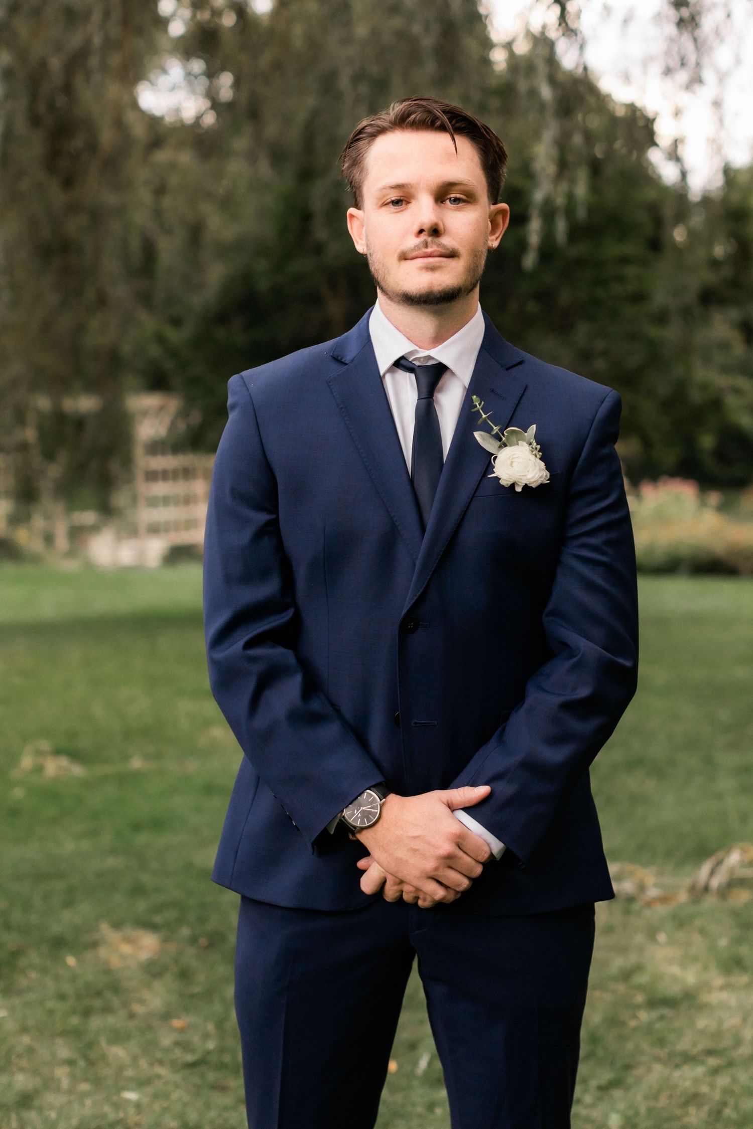 Formal portrait of groom in navy suit standing in garden setting.