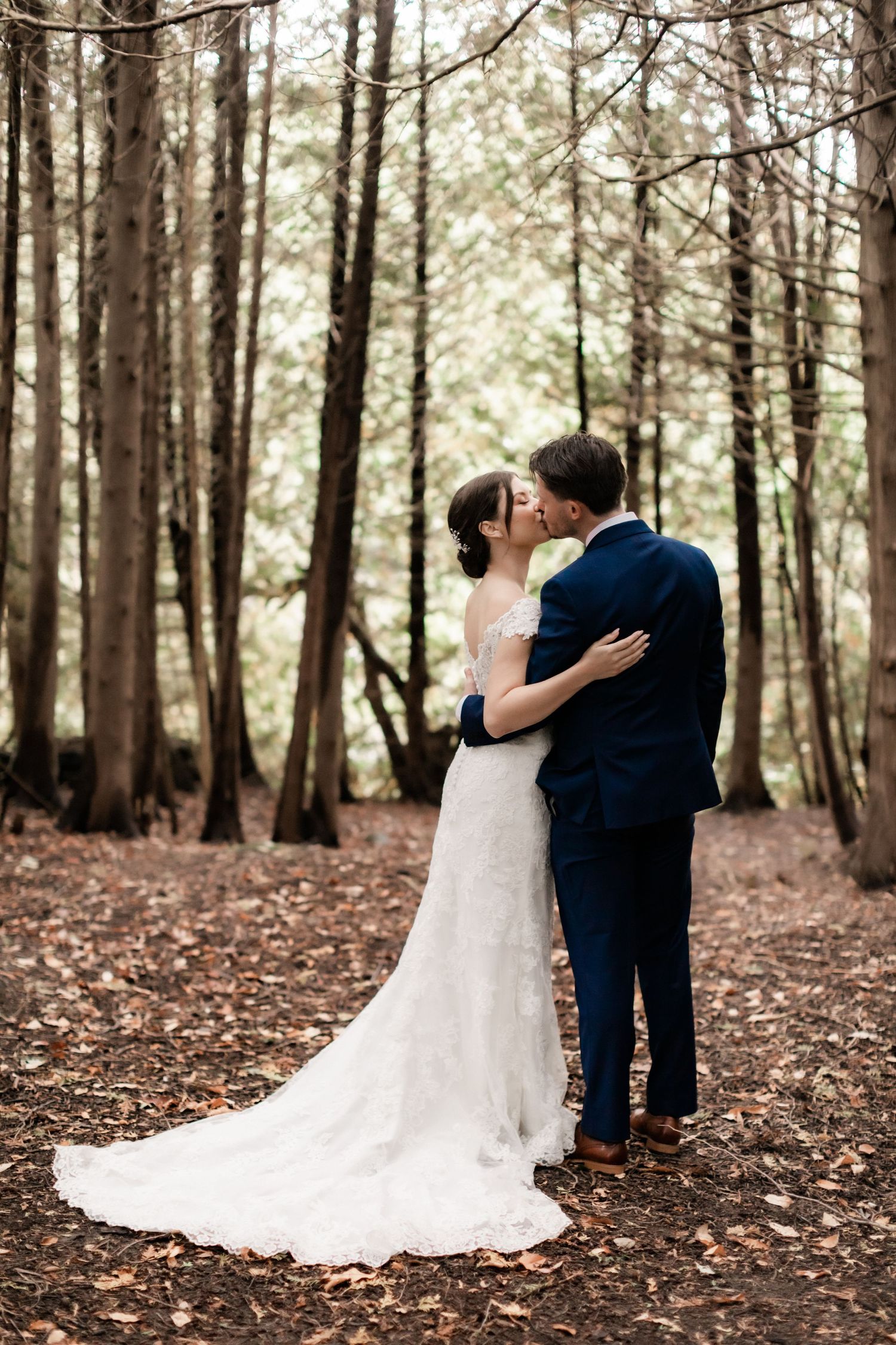 Elegant wedding portrait in forest with bride's lace train trailing on leaf-covered ground.