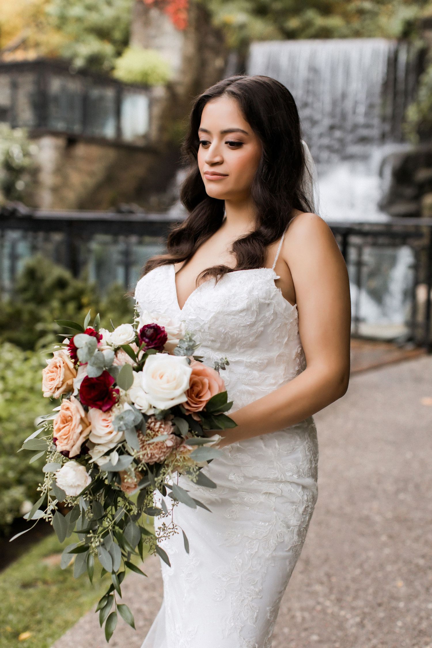 Close-up bridal portrait with cascading bouquet of burgundy and peach roses near waterfall.