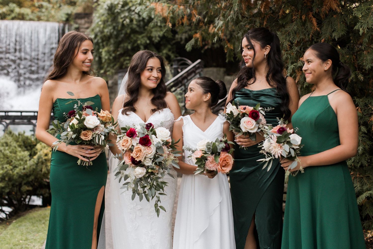 Bridesmaids in elegant green dresses hold floral bouquets while standing together outdoors.