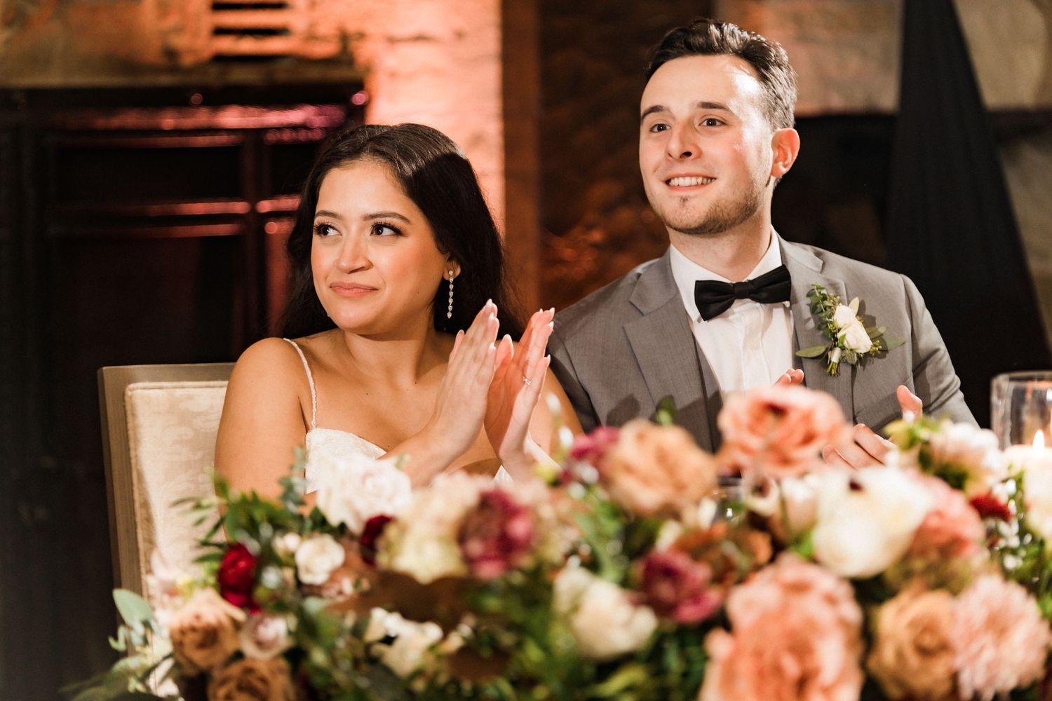 Wedding couple applauding during reception speech with elegant floral centerpiece in foreground.