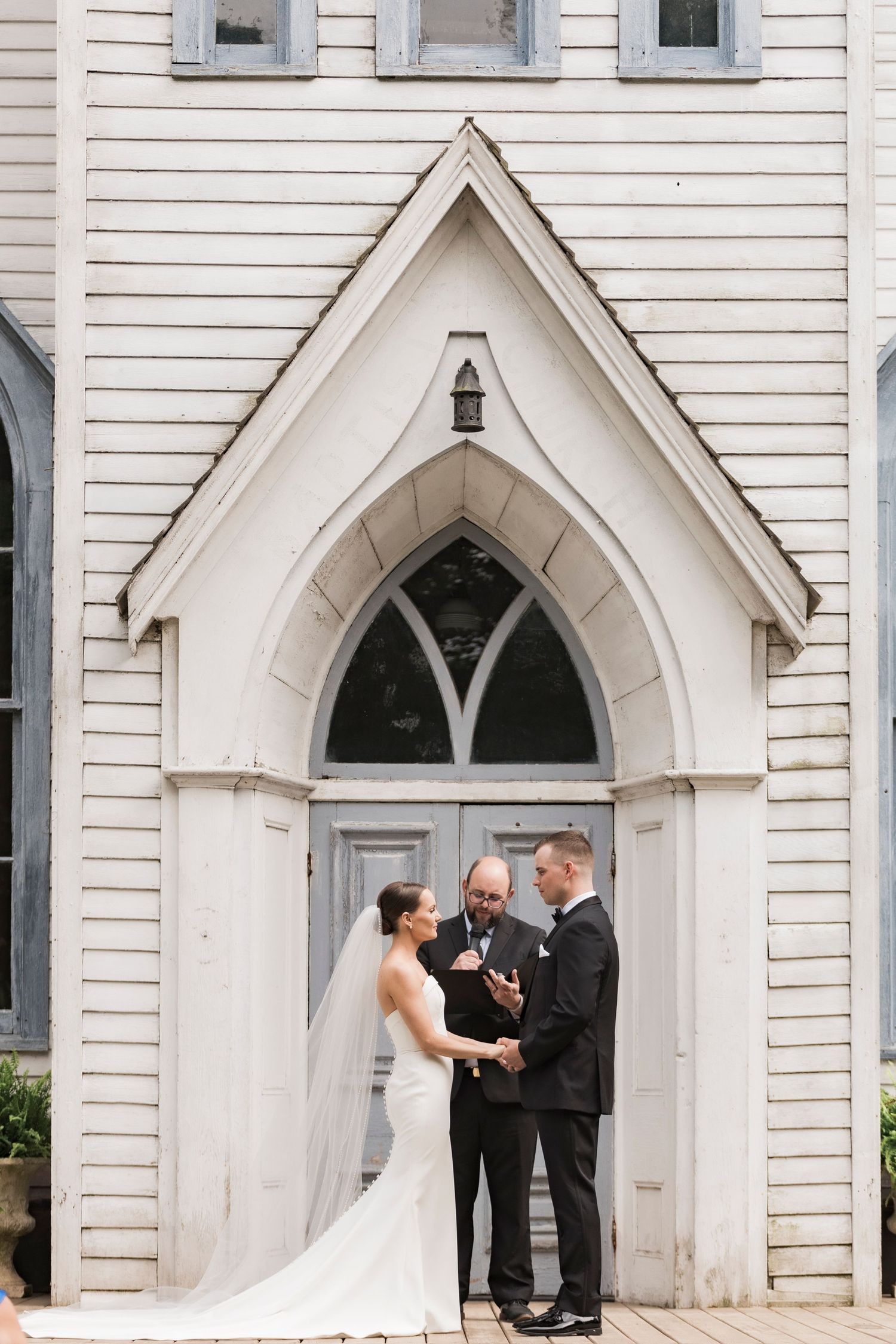 Wedding ceremony takes place in front of a charming white gothic church doorway with pointed arch architecture.