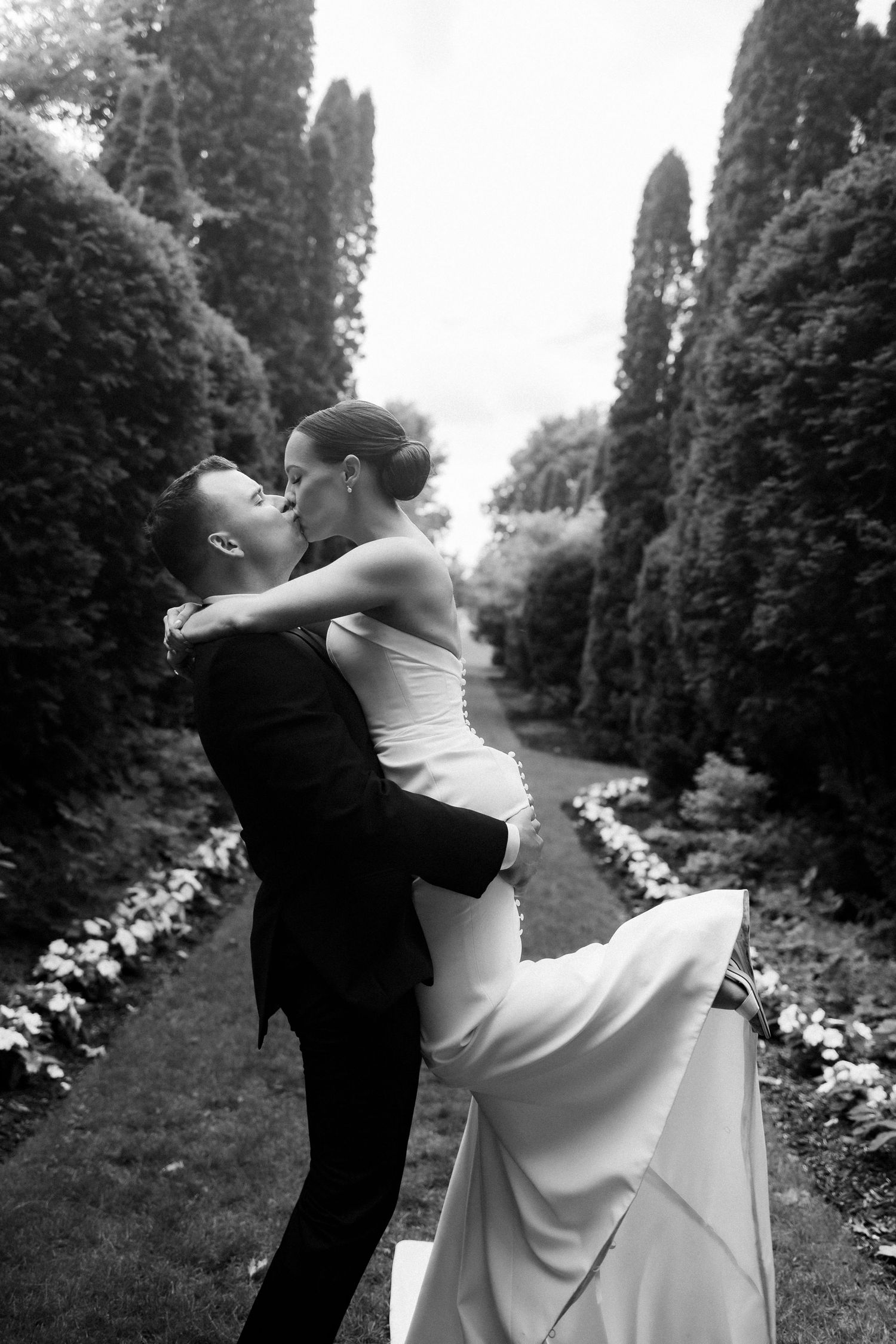 Romantic black and white wedding photo of couple embracing in a garden path lined with tall hedges.