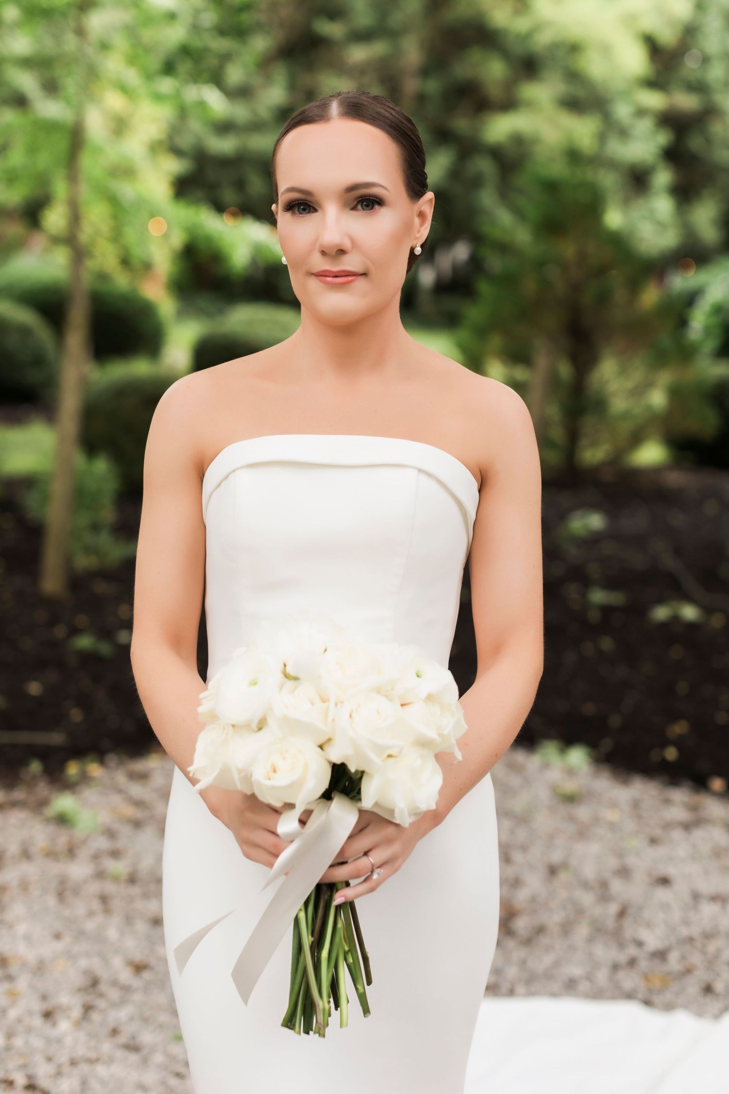 Elegant bride in a strapless white gown holds a classic bouquet of white roses against a garden backdrop.
