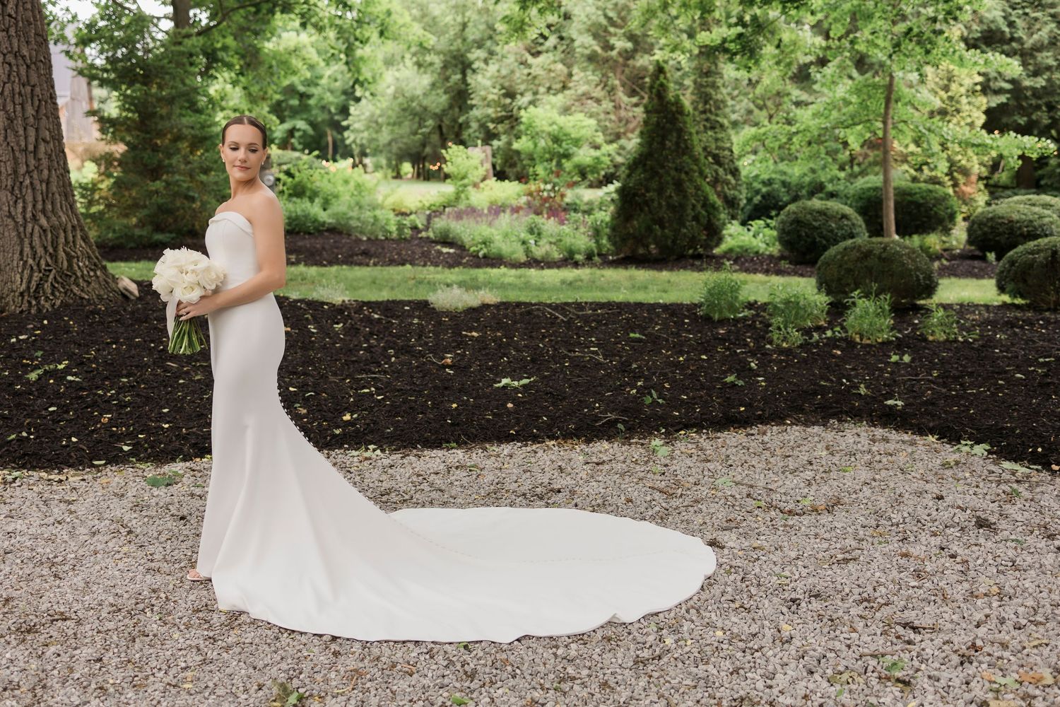 A person in a flowing white wedding dress walks through a lush garden setting with beautiful landscaping and greenery.