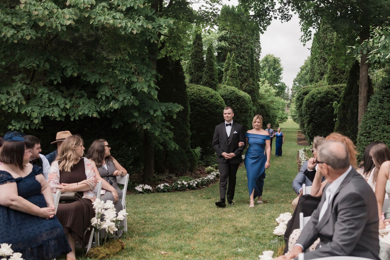 Wedding ceremony outdoors on a lush green lawn with guests seated while a couple walks down the garden aisle.