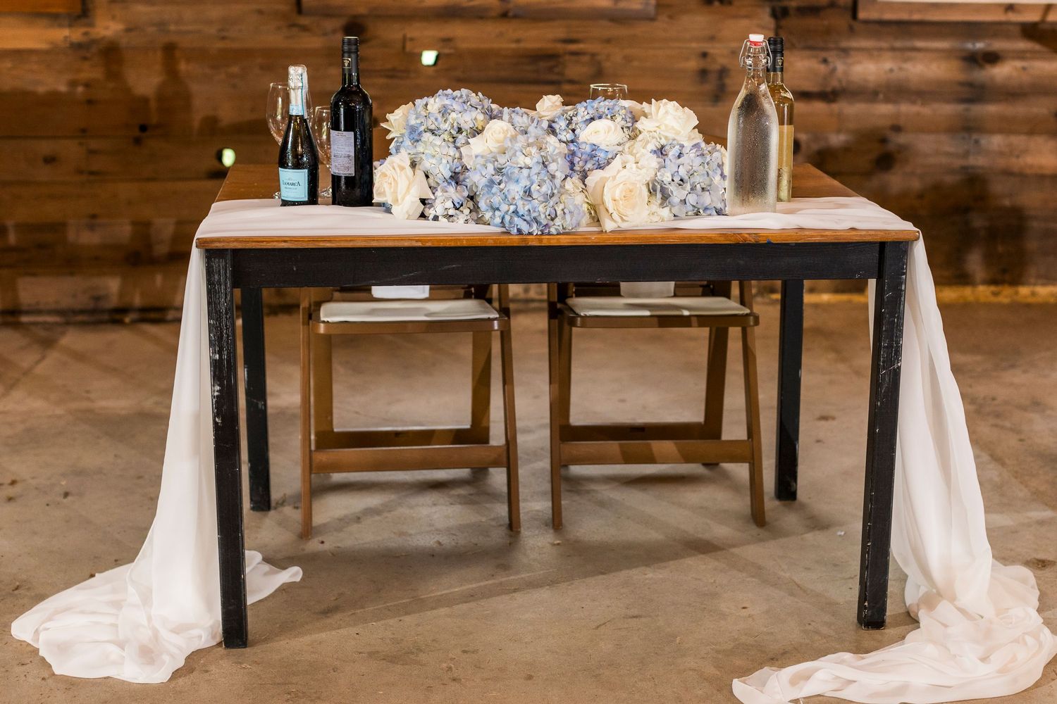 Two trestle tables draped with ivory linens create an elegant wedding display in a rustic wooden barn setting.