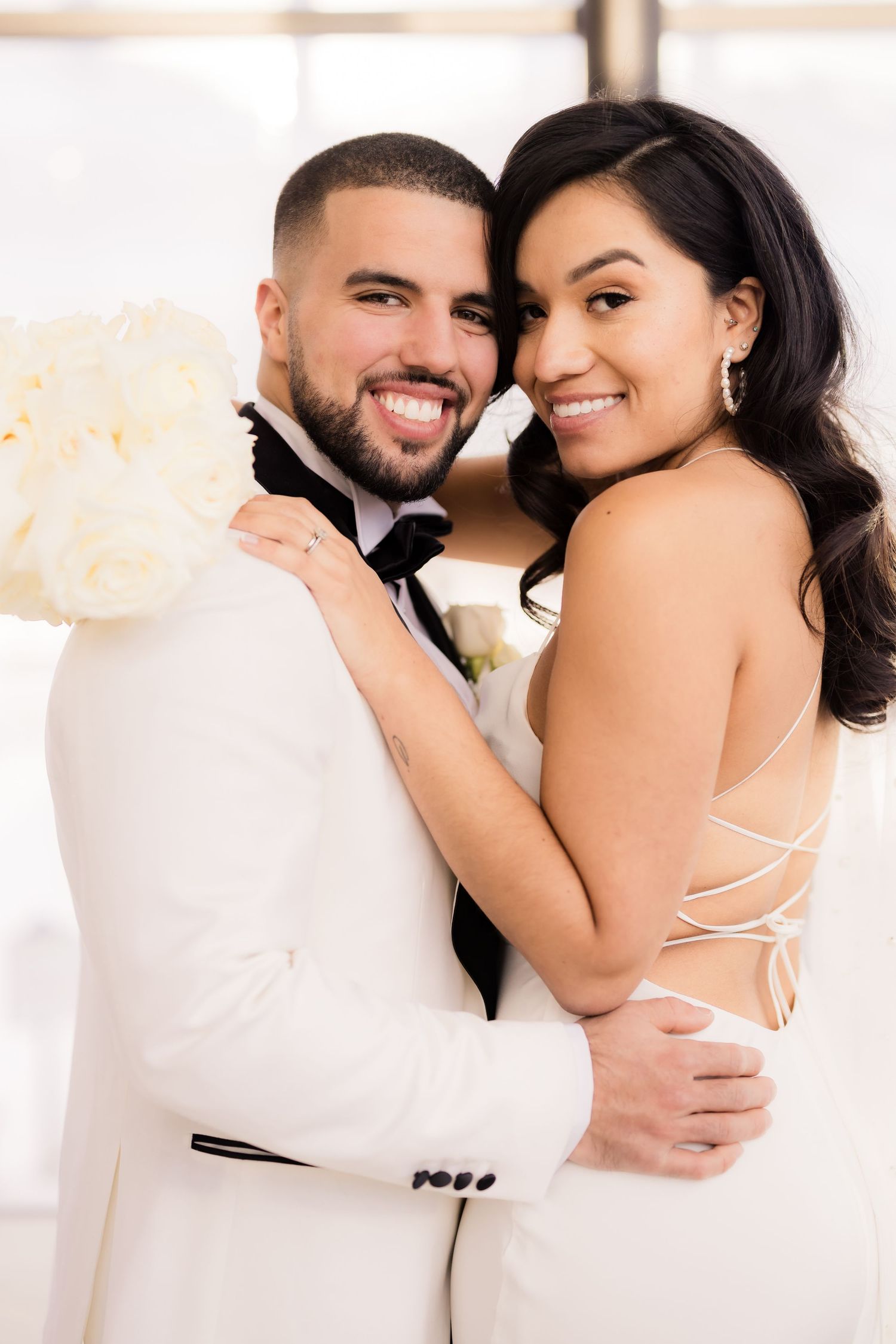 A newlywed couple shares an intimate moment in white attire with a bouquet of cream-colored flowers.