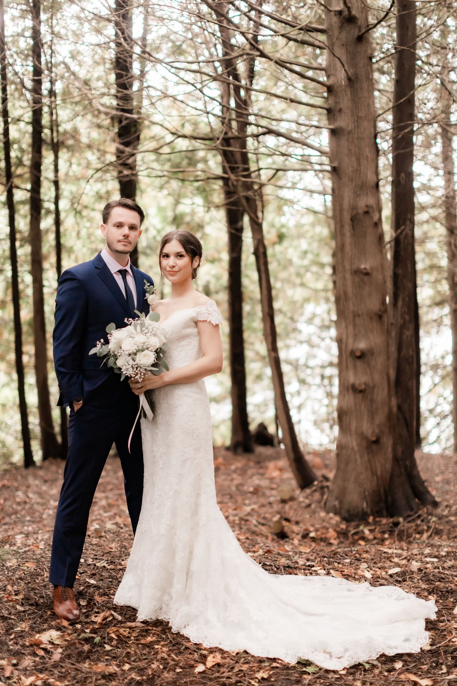 Elegant wedding couple standing in a pine forest with bride's lace dress trailing behind.