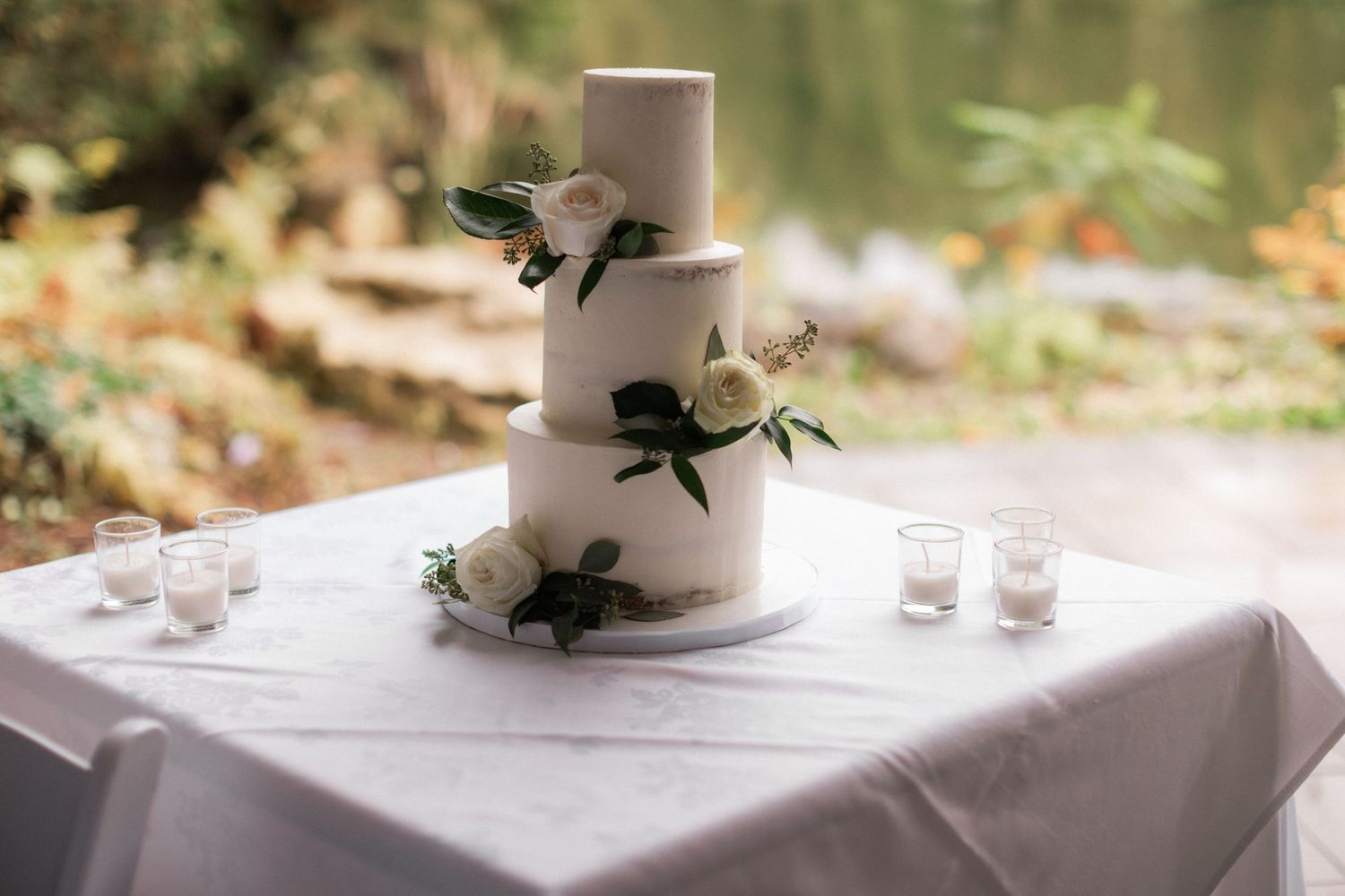 An elegant three-tier white wedding cake decorated with fresh greenery and flowers on a table.