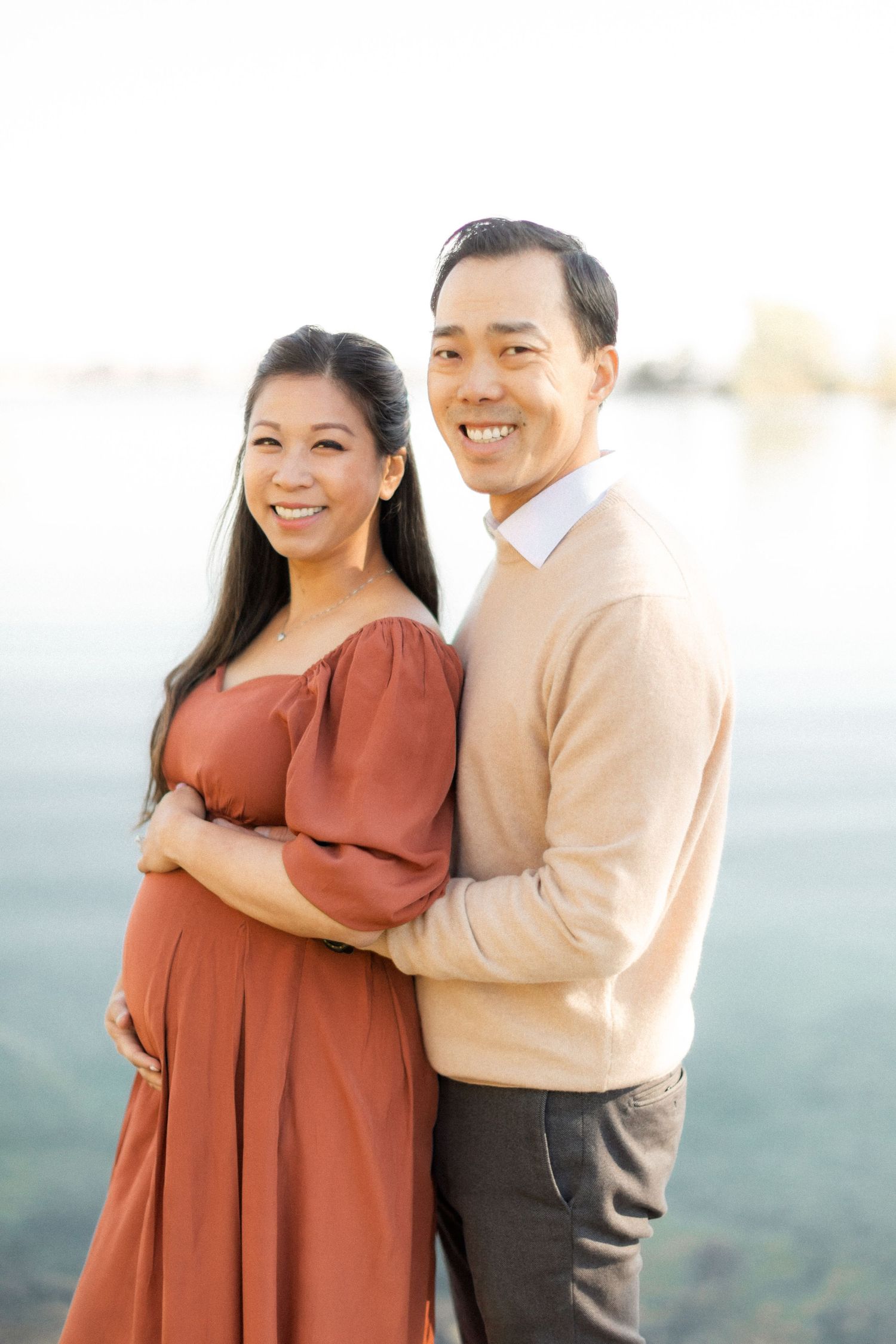Joyful maternity portrait of couple by lake in coordinating autumn colors.