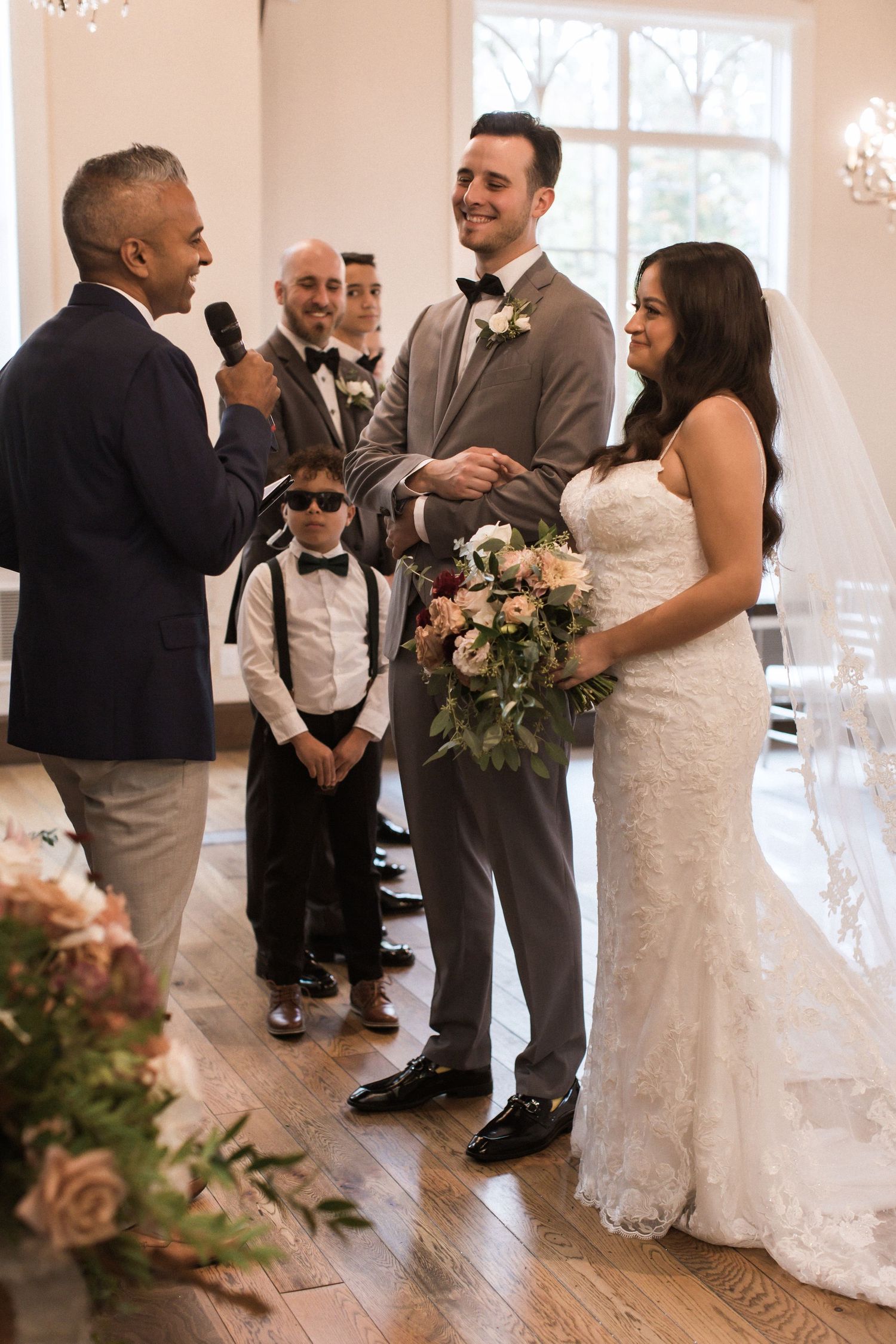 A wedding ceremony in progress with a couple and officiant standing in a bright white room with elegant decor.