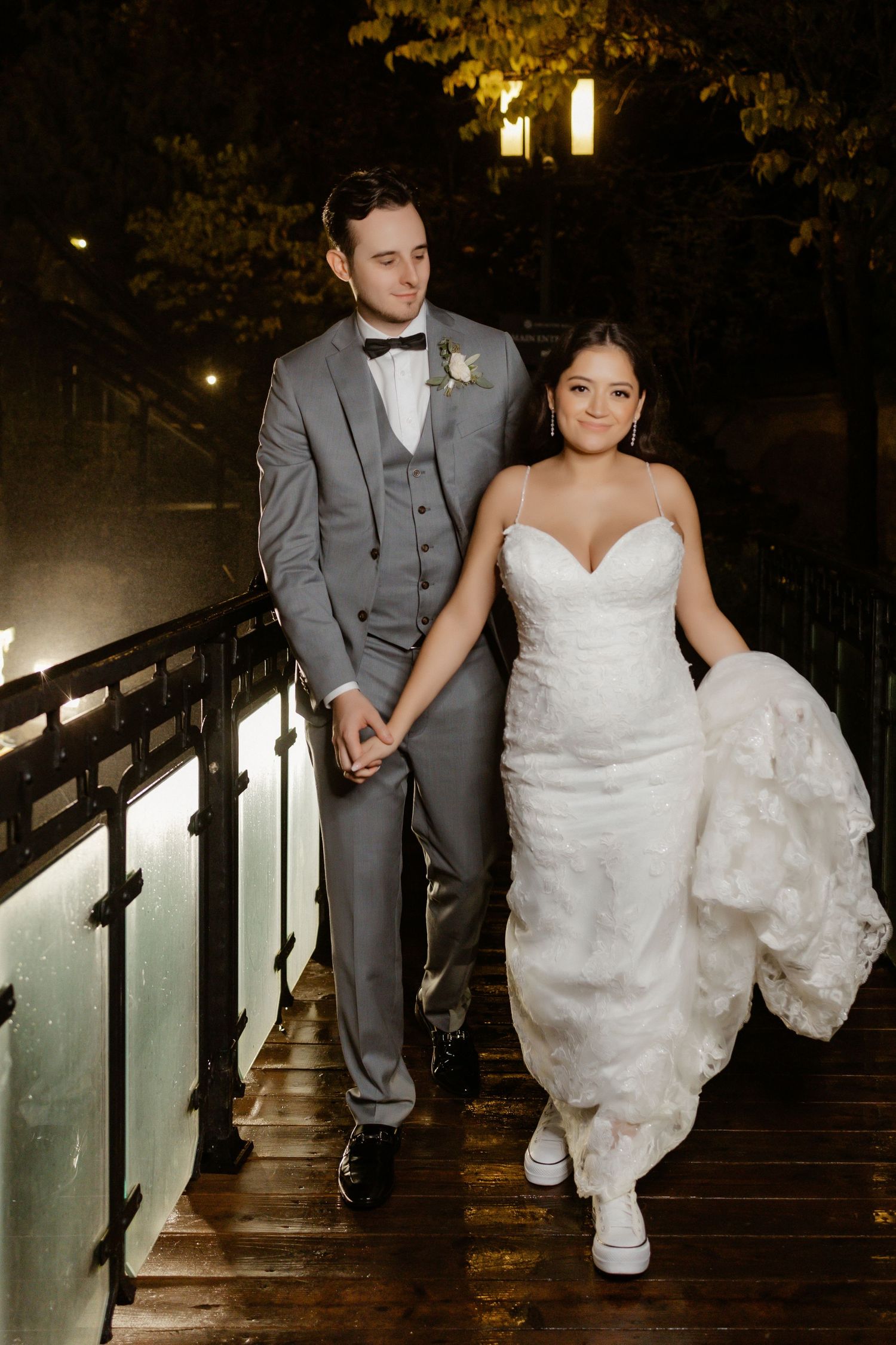 A couple in wedding attire walks along a dimly lit wooden boardwalk at night.