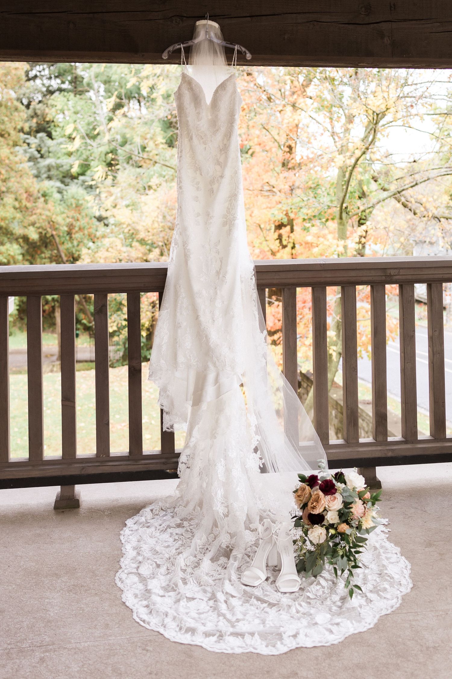 Elegant white lace wedding dress hanging on display on a covered porch with fall foliage in background.