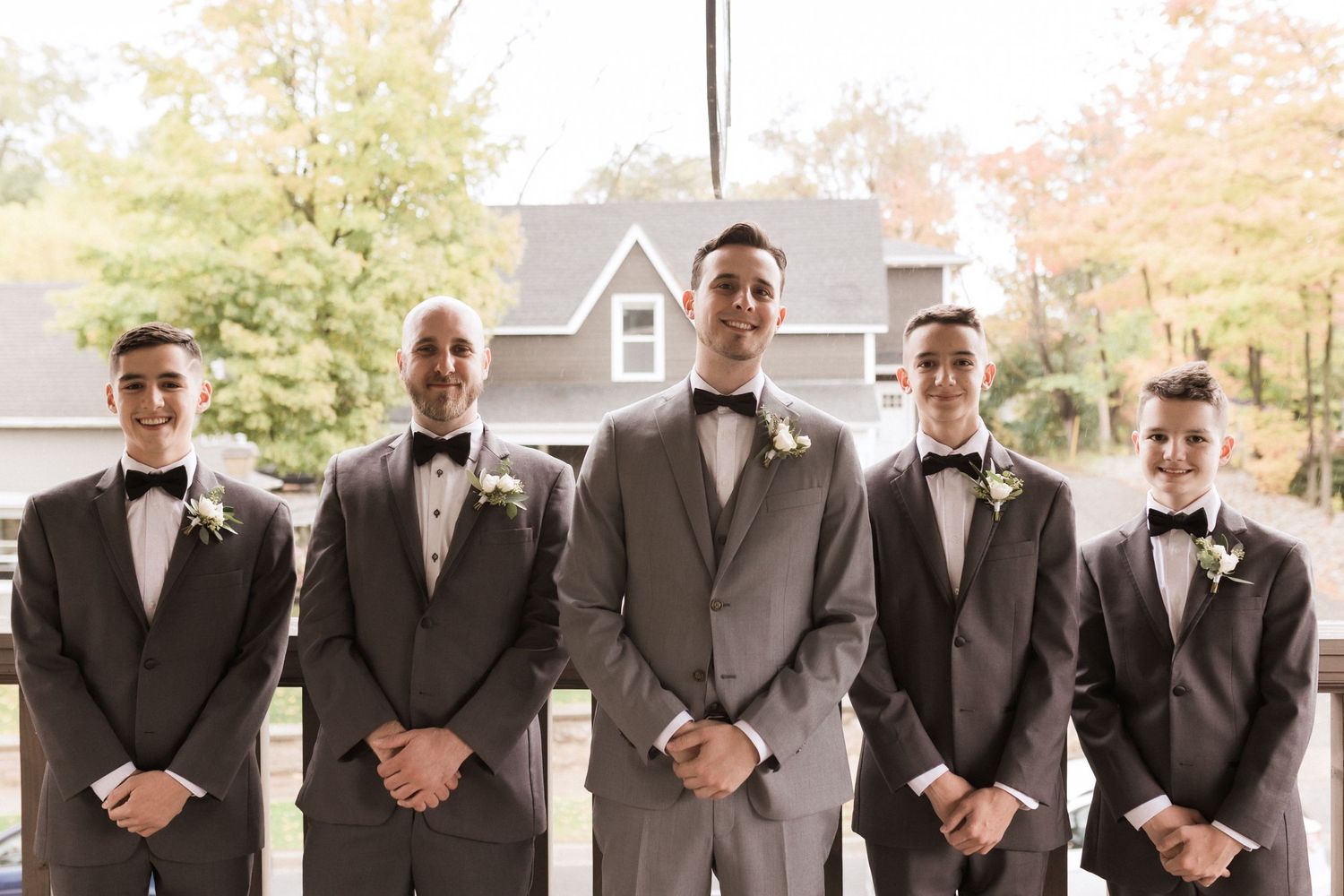 Wedding party of five groomsmen in formal suits pose together outdoors.