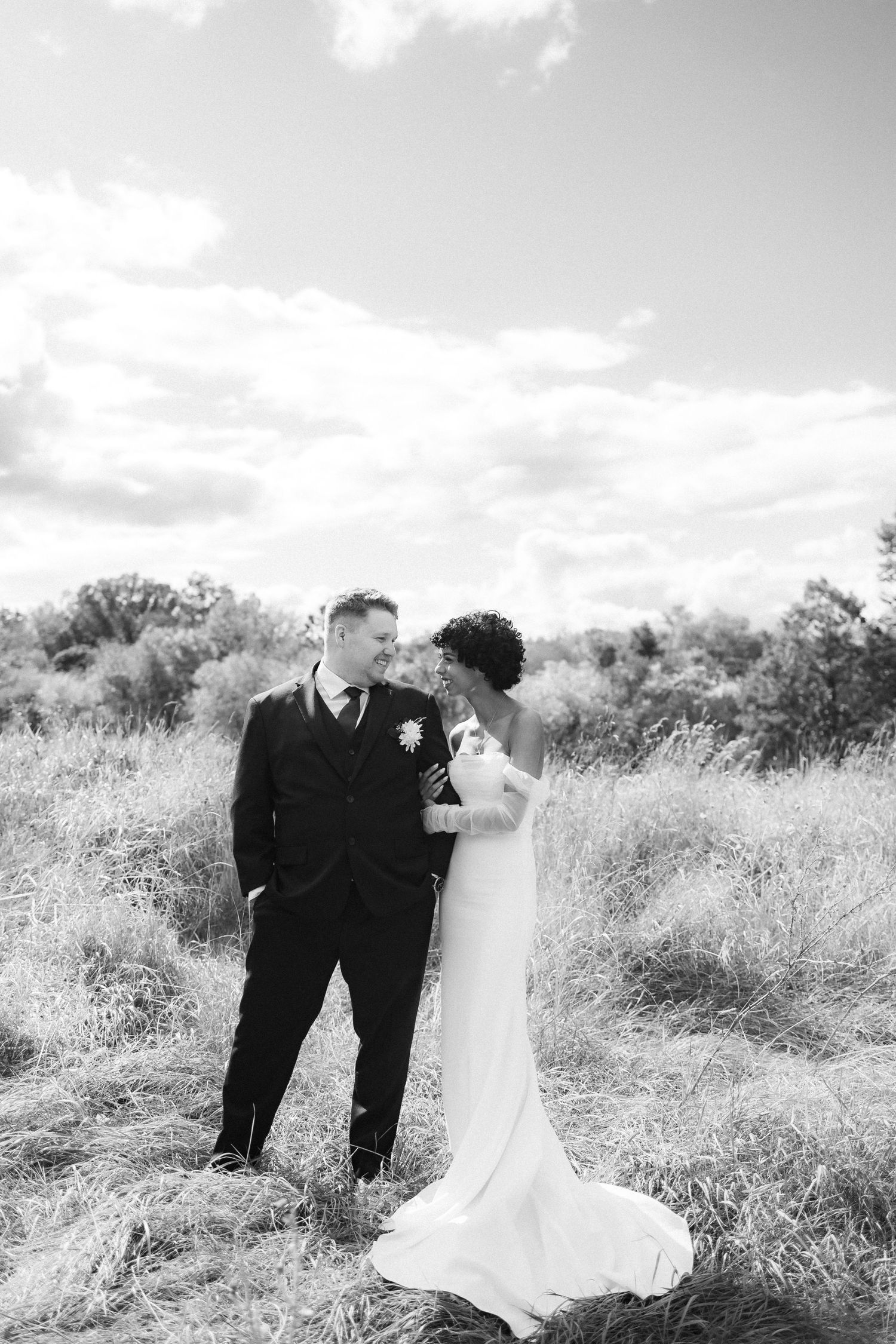 Black and white photo of a wedding couple standing in a meadow under cloudy skies.