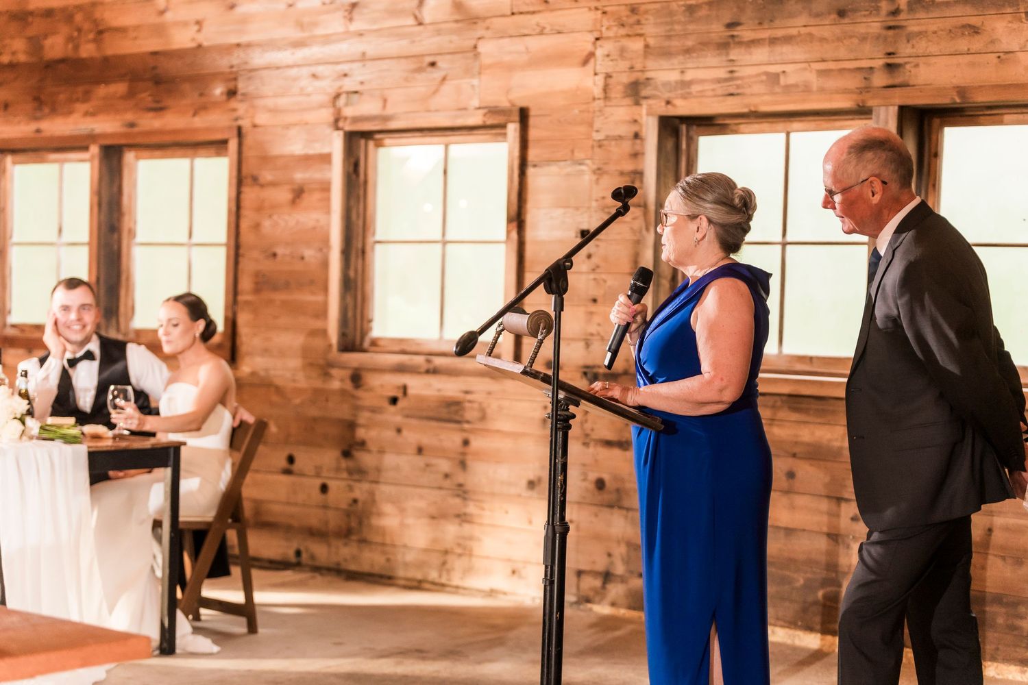 Two speakers deliver remarks at a rustic wooden barn venue during a wedding reception.