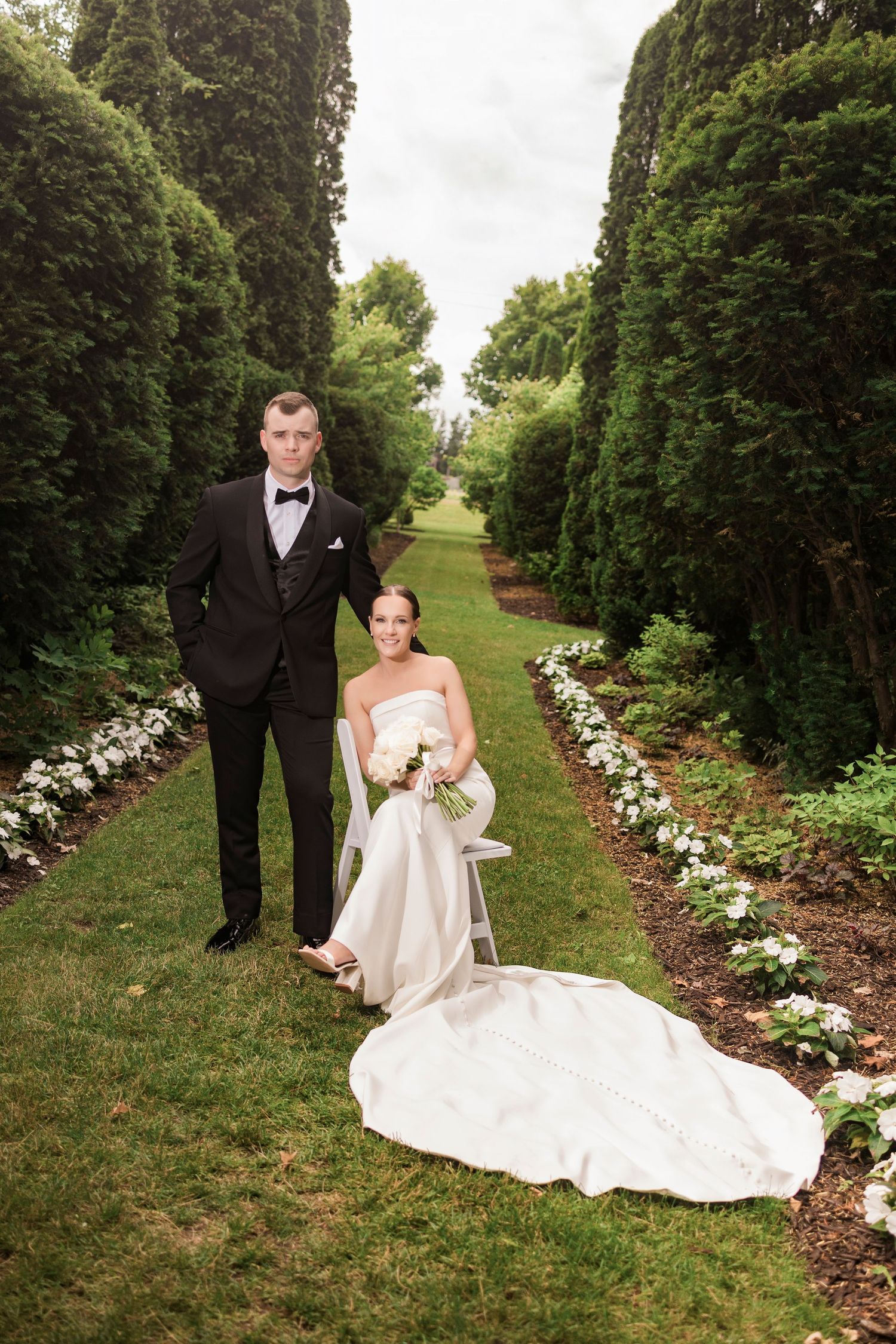 Wedding portrait of couple posing on garden path with flowing white dress train and tall cypress trees.