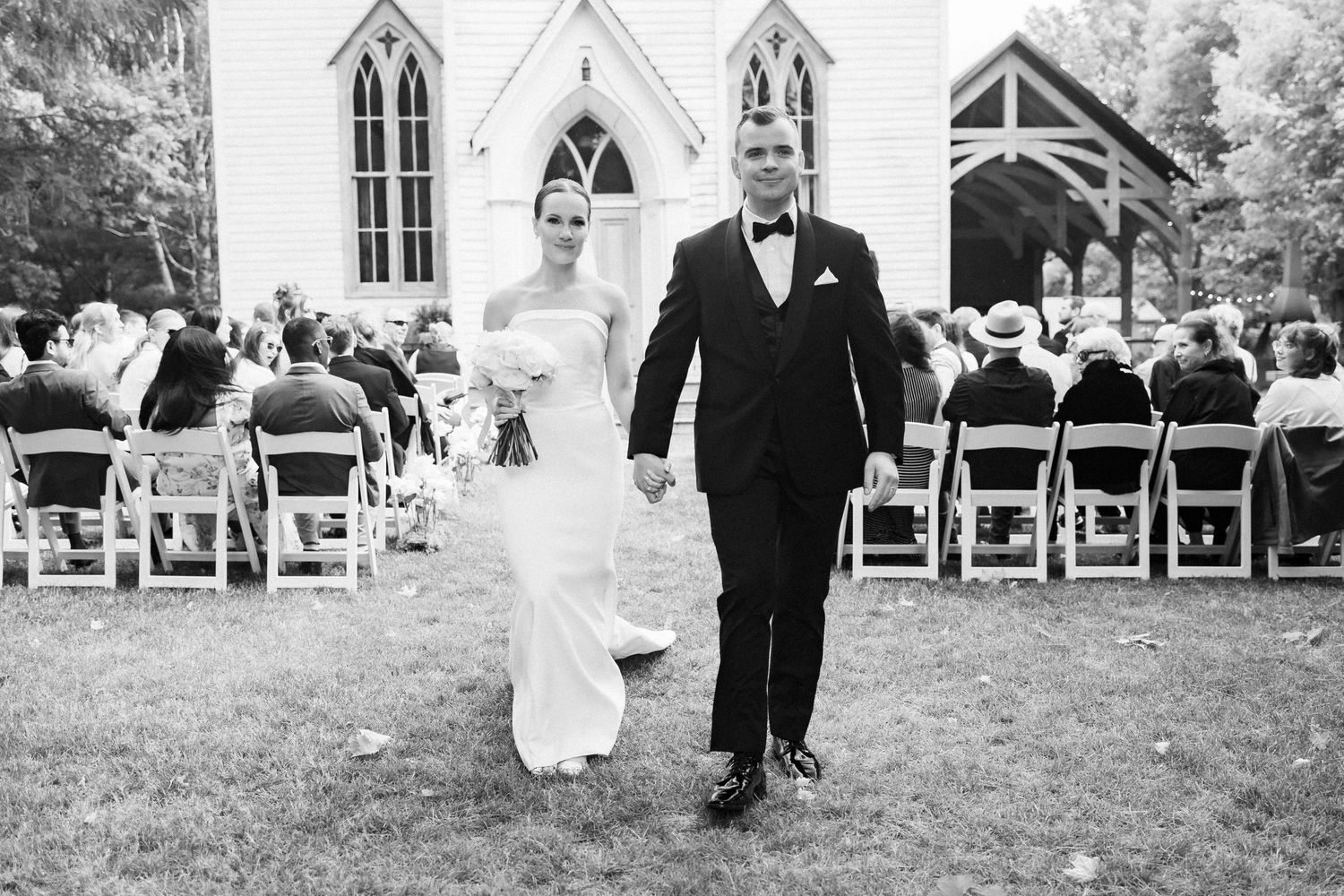 Wedding ceremony guests observing the outdoor celebration against a backdrop of a white church with Gothic windows.