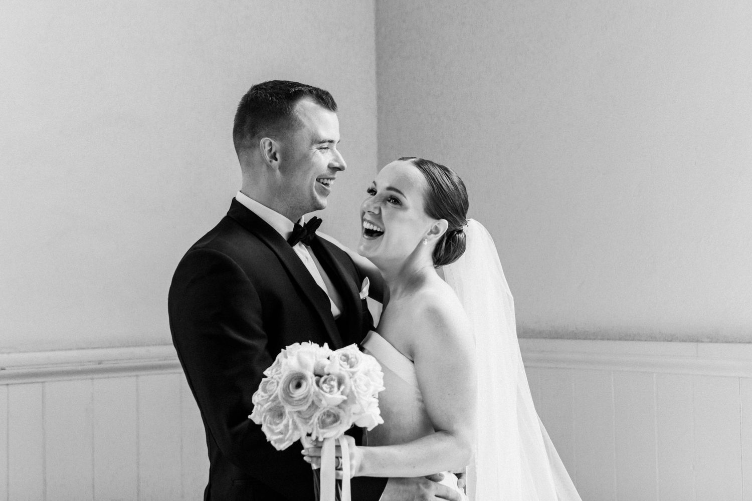 A joyful black and white wedding photo showing a couple laughing together while holding a white floral bouquet.