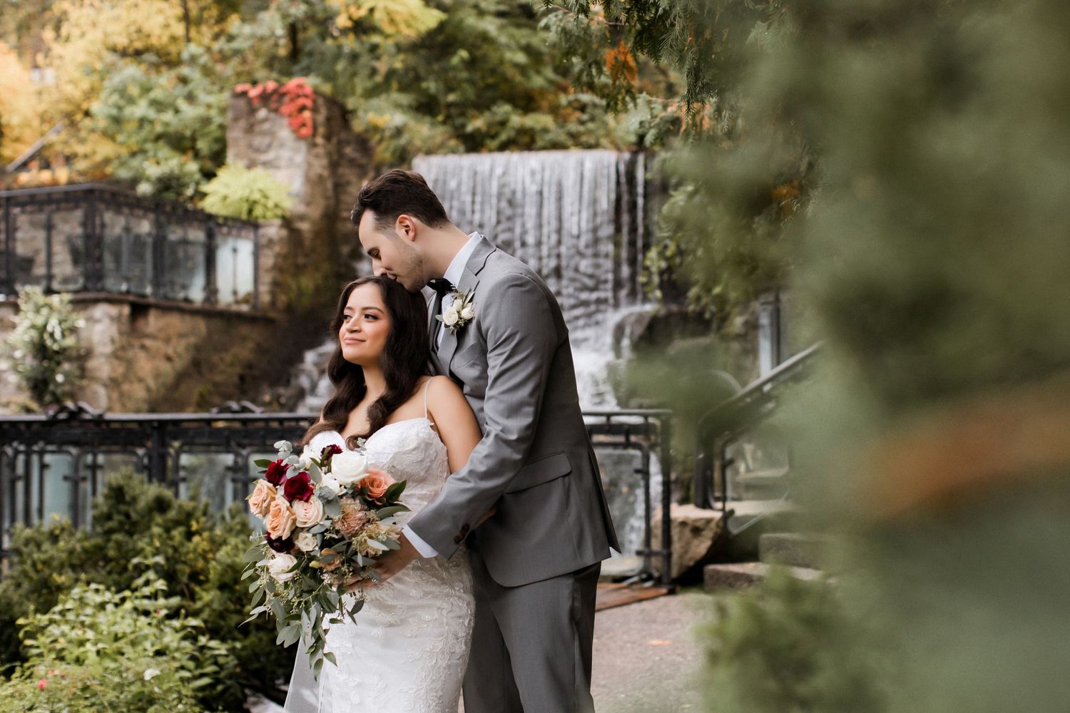 Tender moment between newlyweds in garden setting with waterfall backdrop.