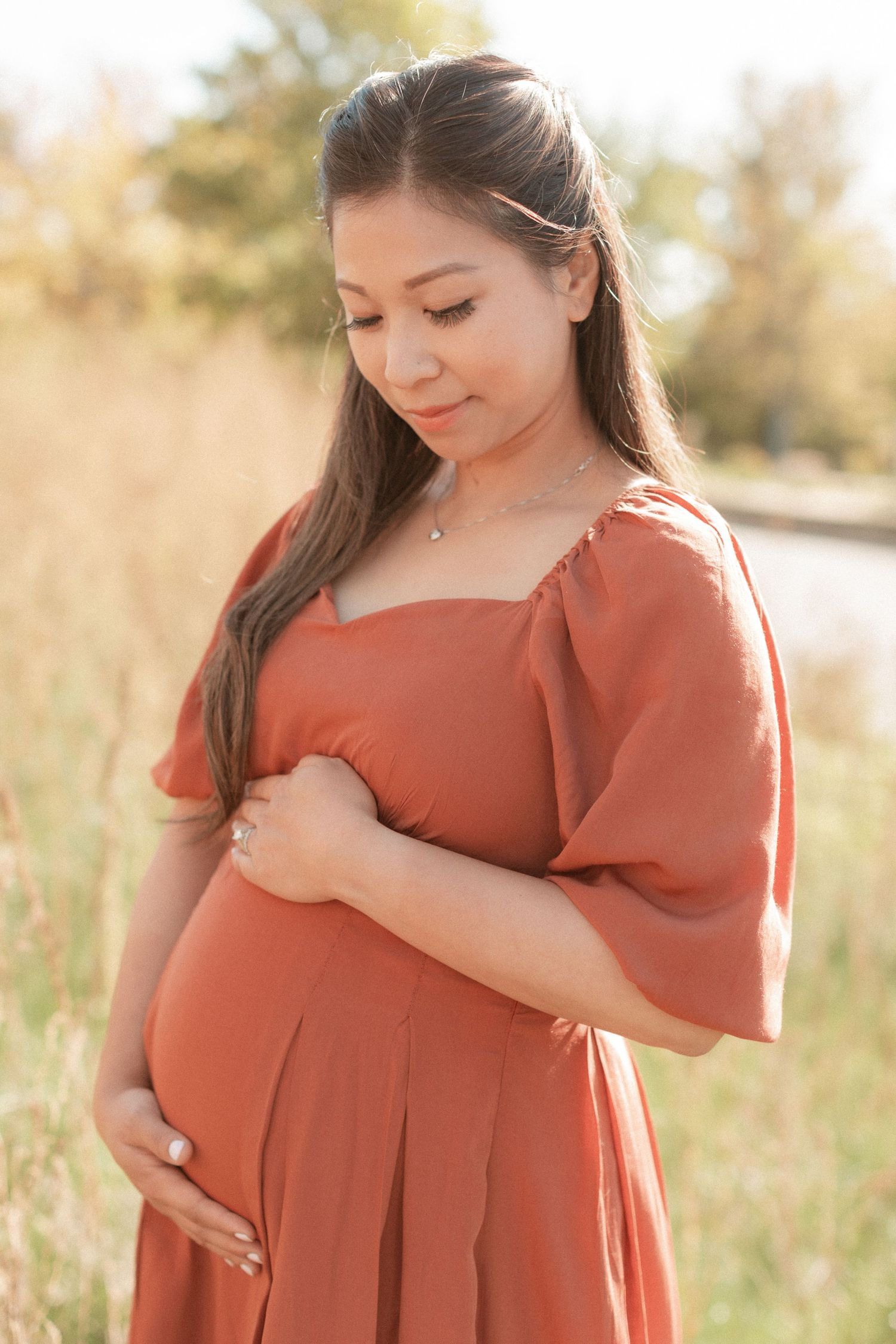 Expectant mother in a rust-colored dress stands in a sunlit field holding her baby bump.