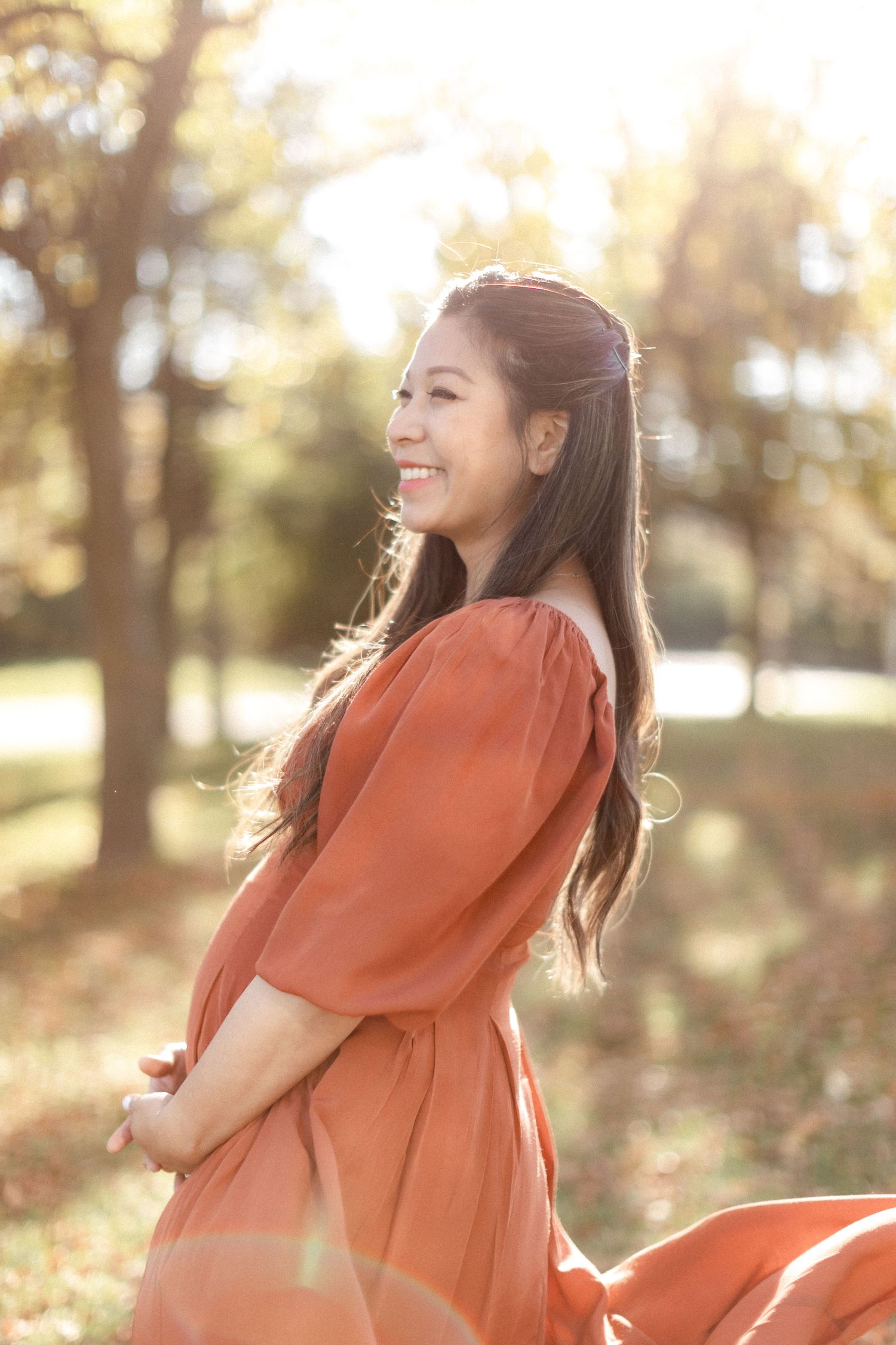 Radiant maternity portrait of woman in rust dress with sunlight creating a warm glow.