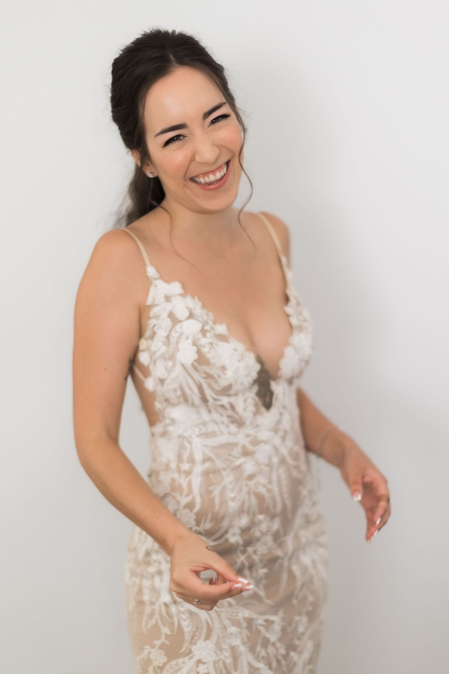 Woman in white lace gown laughs joyfully against plain background.