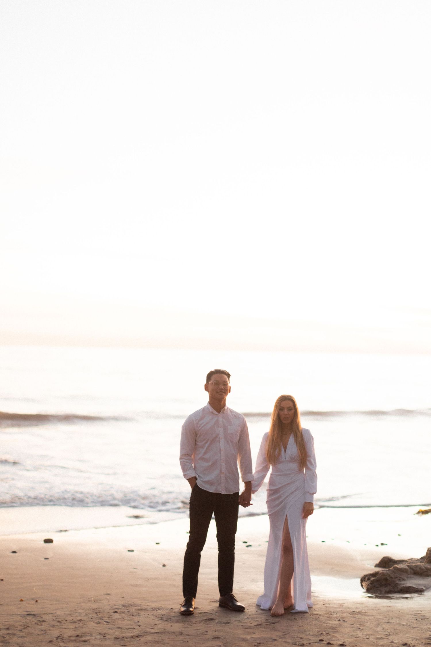 Couple holding hands while walking on beach at sunset in formal white attire.