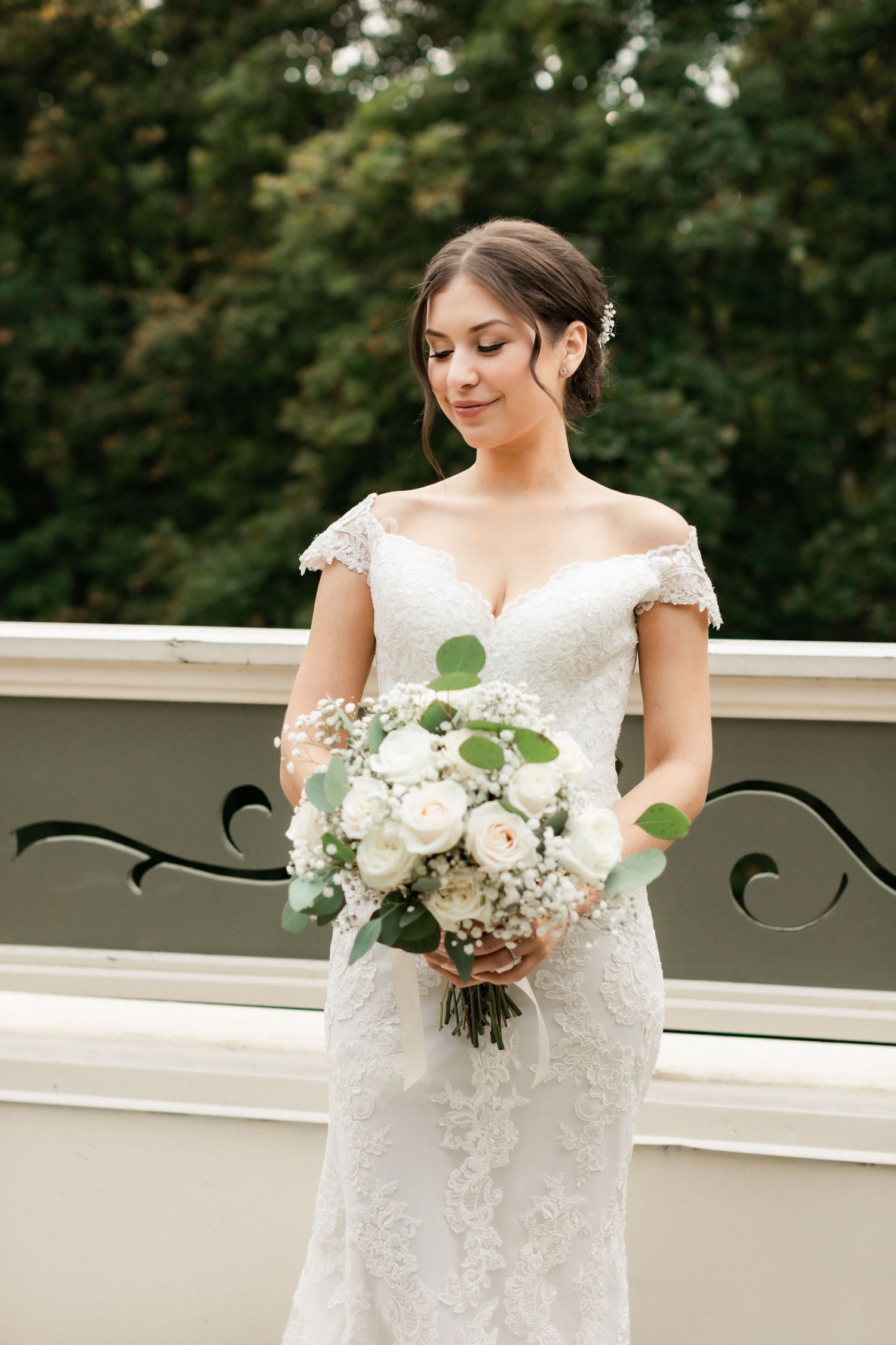 A bride in a lace wedding dress holding a white floral bouquet on a balcony.