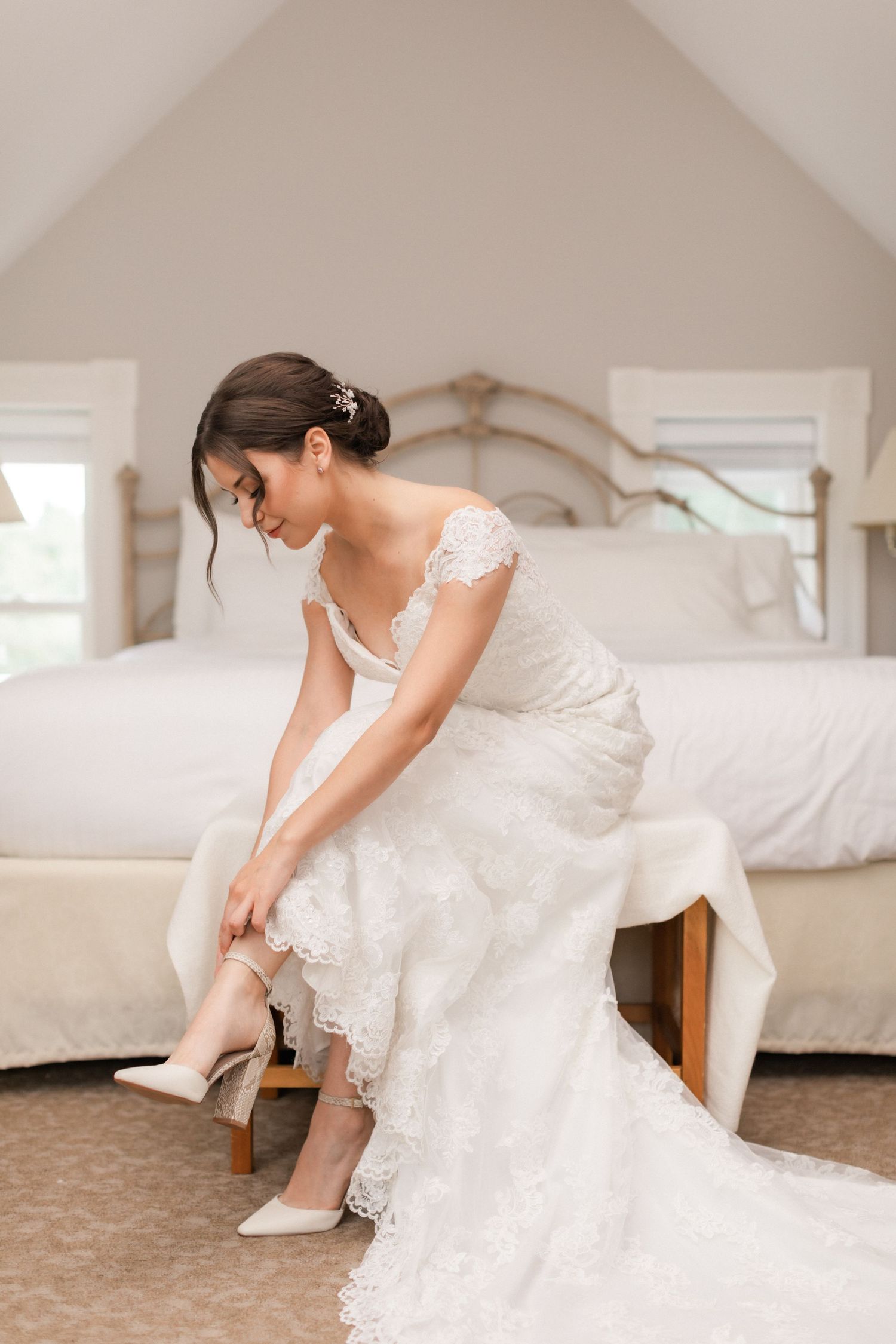Bride puts on wedding shoes while sitting on bench in elegant lace gown.