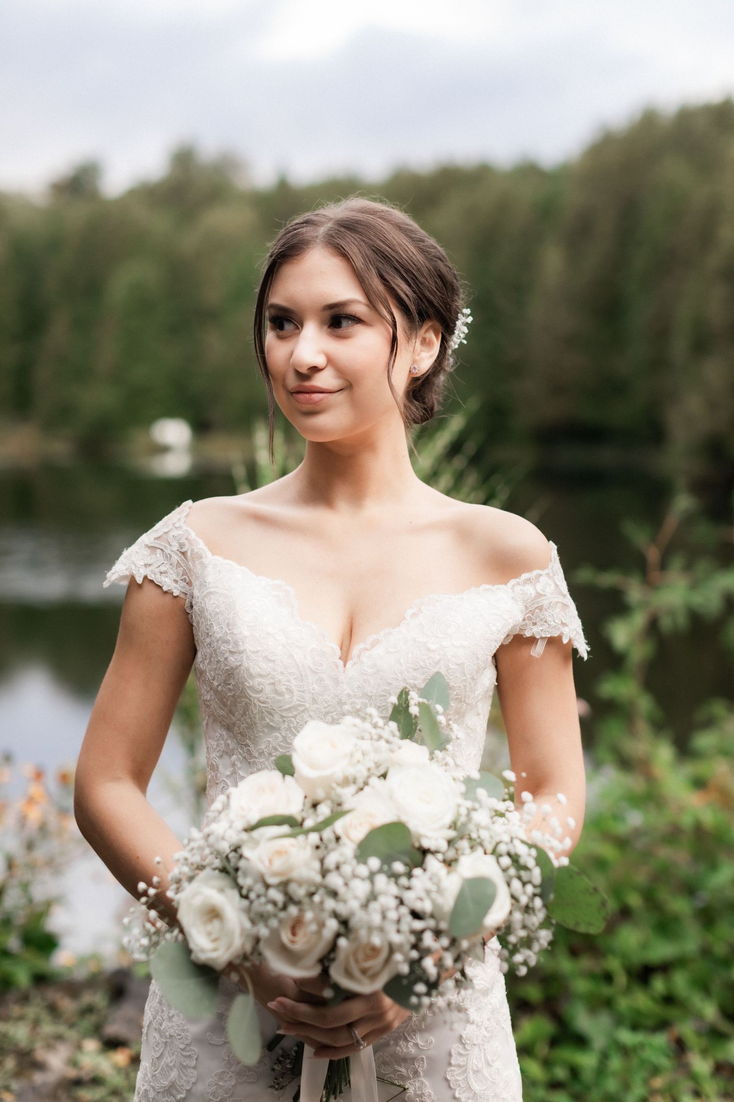 A bridal portrait showcasing an elegant lace dress and white rose bouquet against a lakeside backdrop.