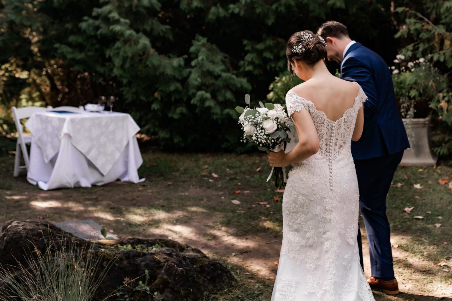Back view of couple in wedding attire standing beside decorated ceremony table outdoors.