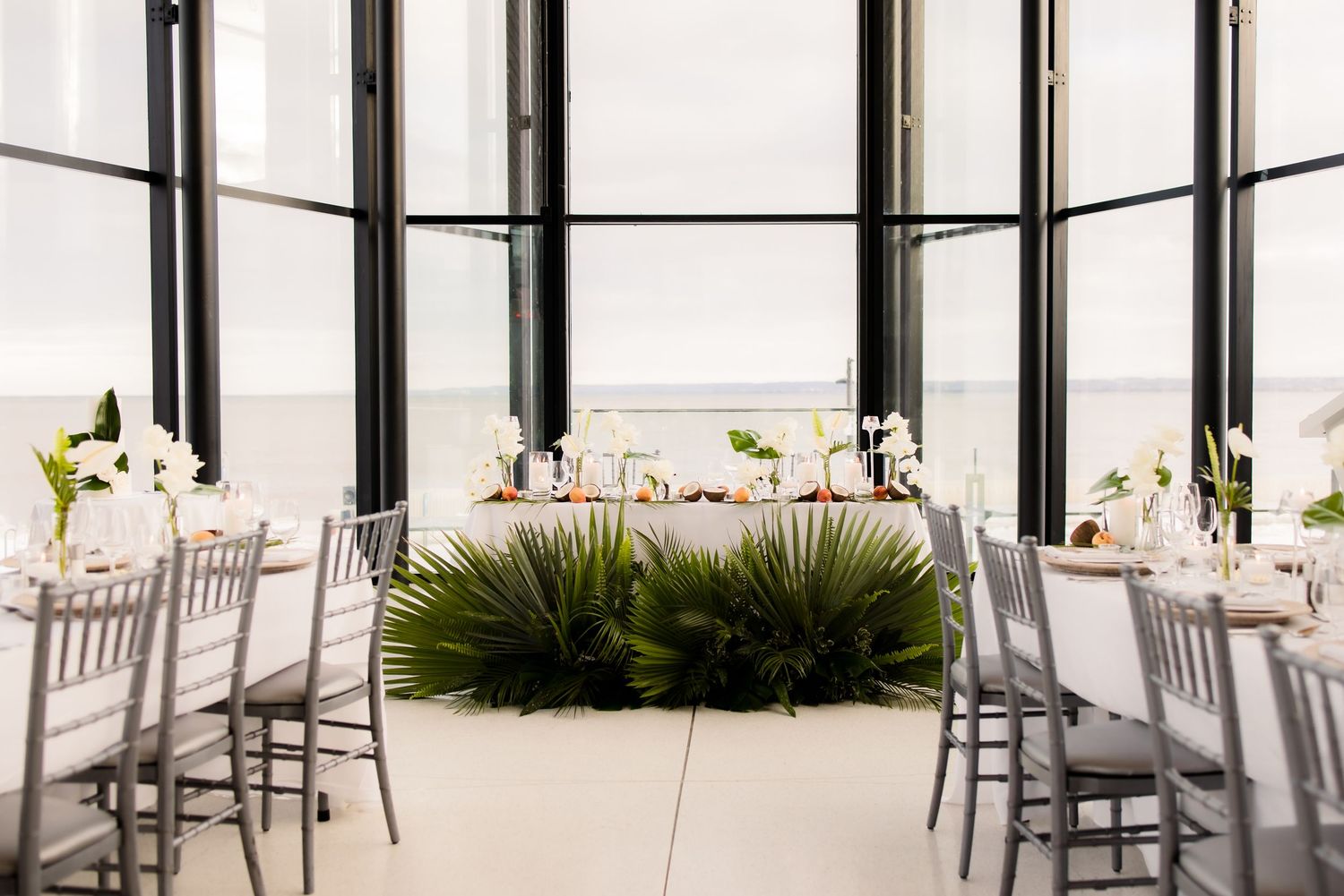 Modern wedding reception setup with tropical greenery centerpieces and silver chiavari chairs in glass-walled venue.