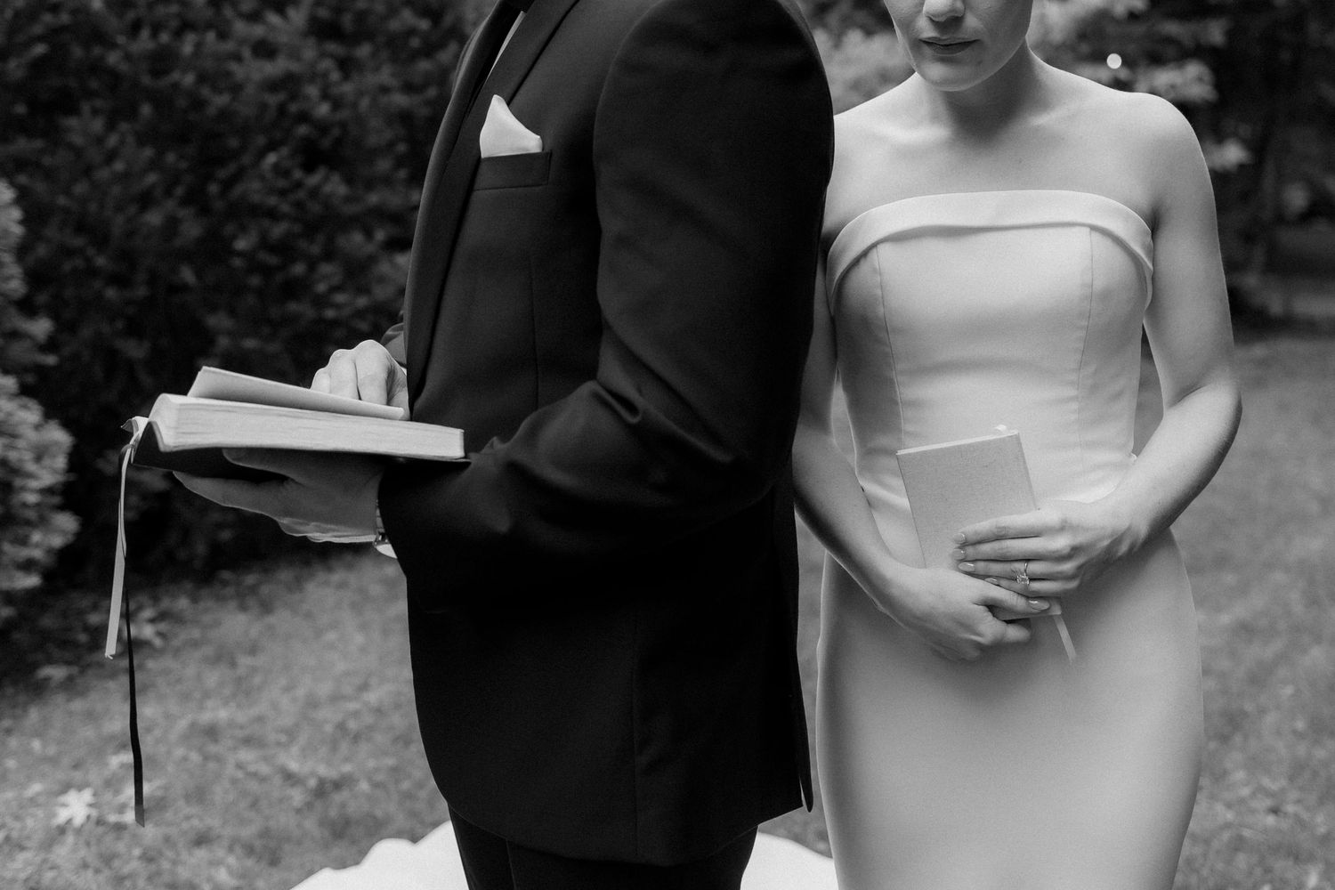 A black and white photo of a bride and groom standing back to back holding books in a garden setting.