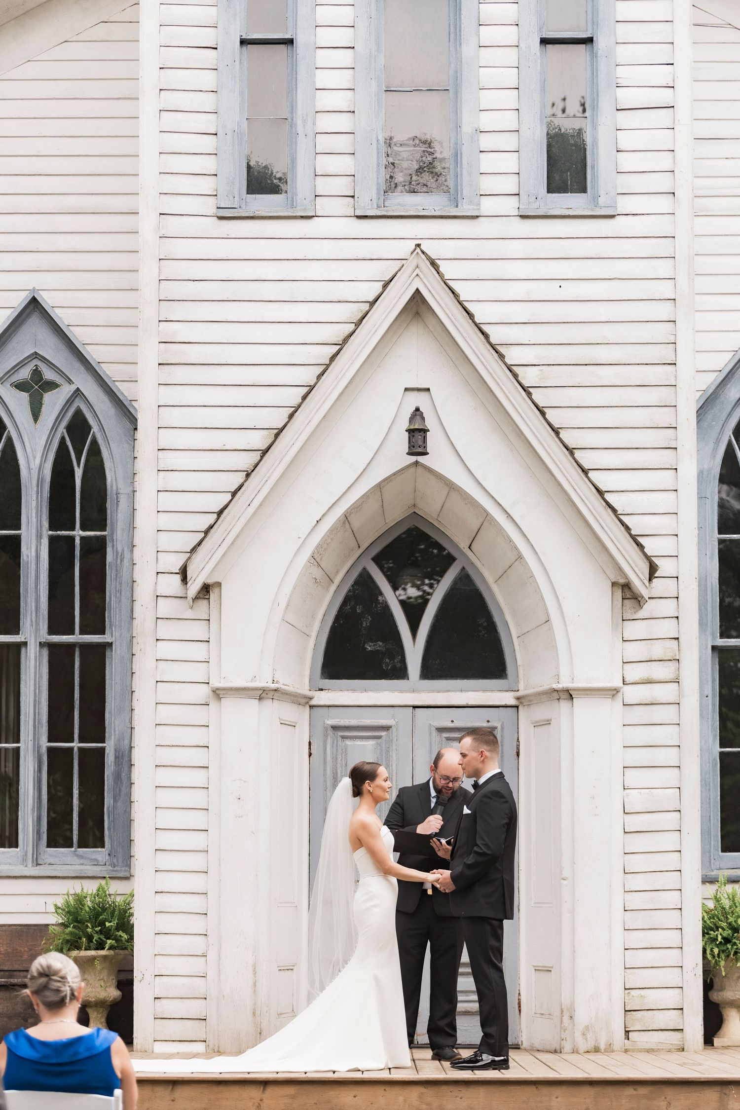 A romantic wedding ceremony takes place in the doorway of a charming white Gothic revival church with arched windows.