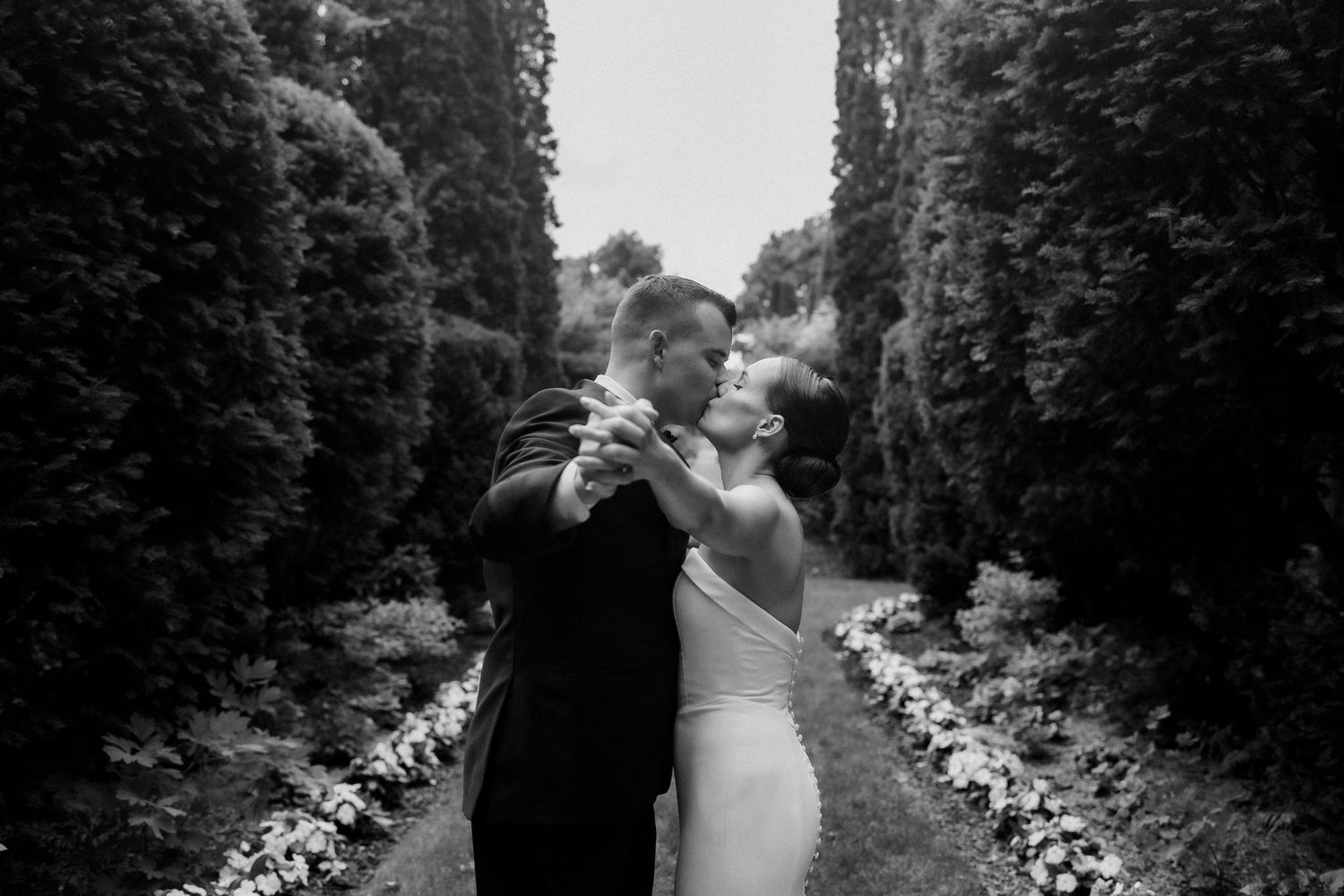 A couple shares an intimate dance moment on a garden path flanked by tall hedges in black and white.