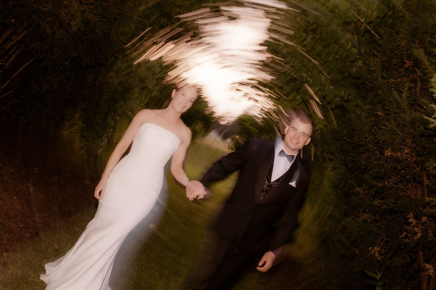 Two figures in wedding attire walk together under a darkened natural outdoor tree canopy on a path.