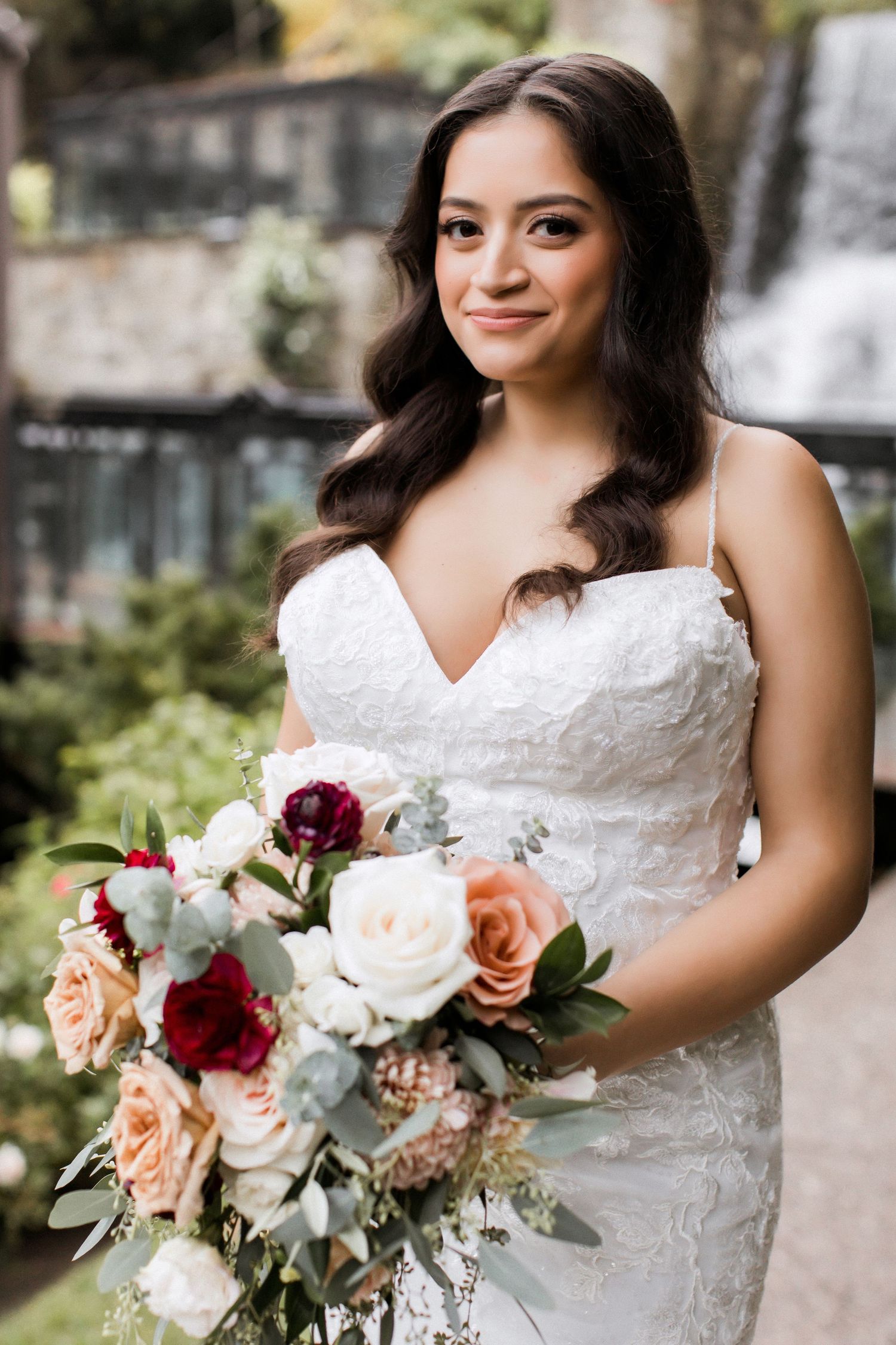 Beautiful bridal portrait with a stunning bouquet featuring burgundy and peach roses against a waterfall backdrop.