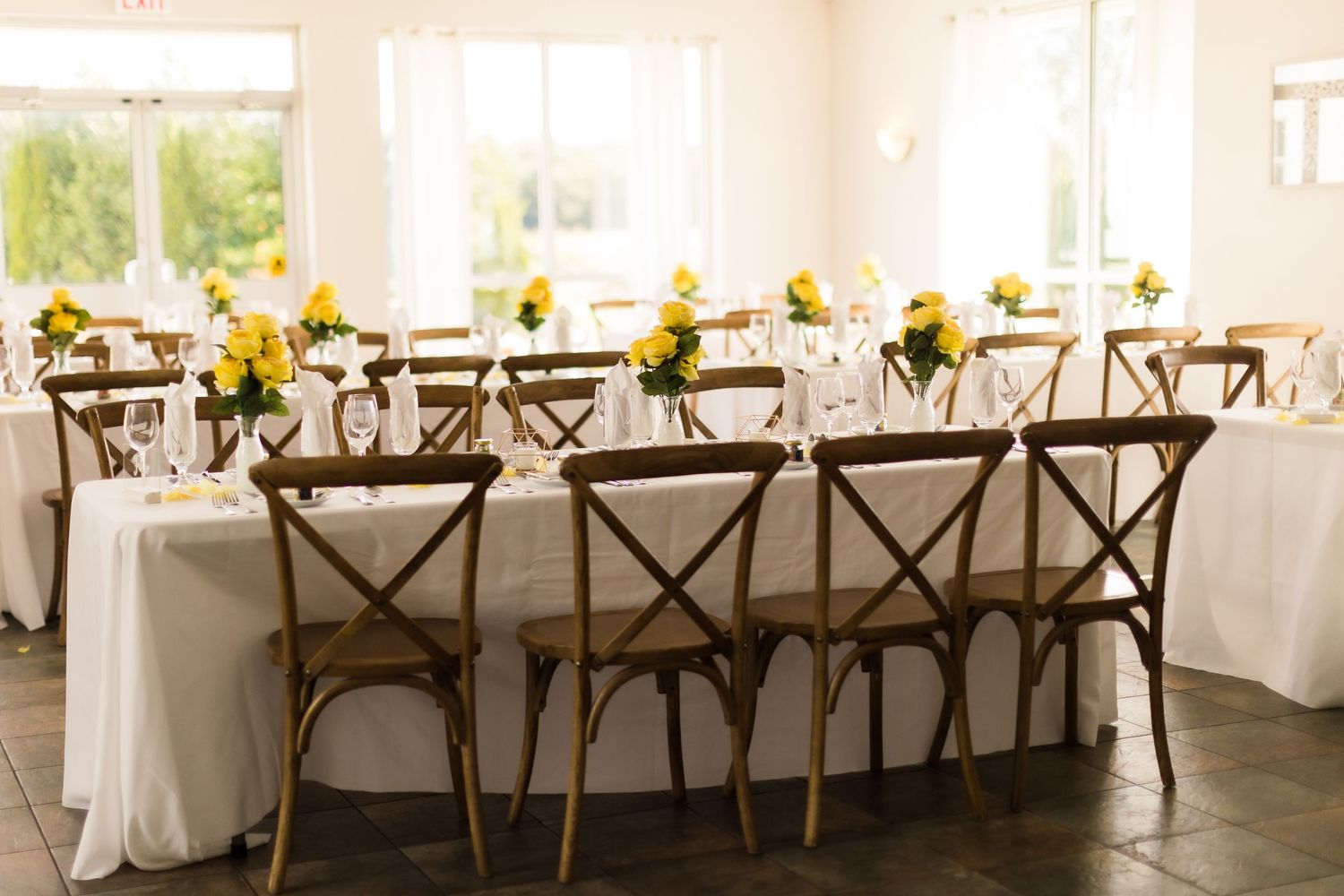 Long banquet table with white linens and crossback chairs decorated with yellow floral arrangements.