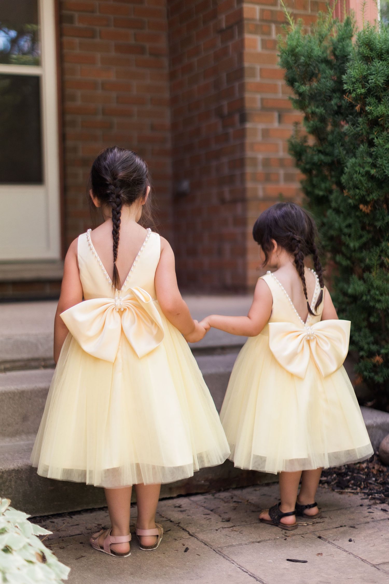 Two young flower girls in matching yellow tulle dresses with large bows walk together.