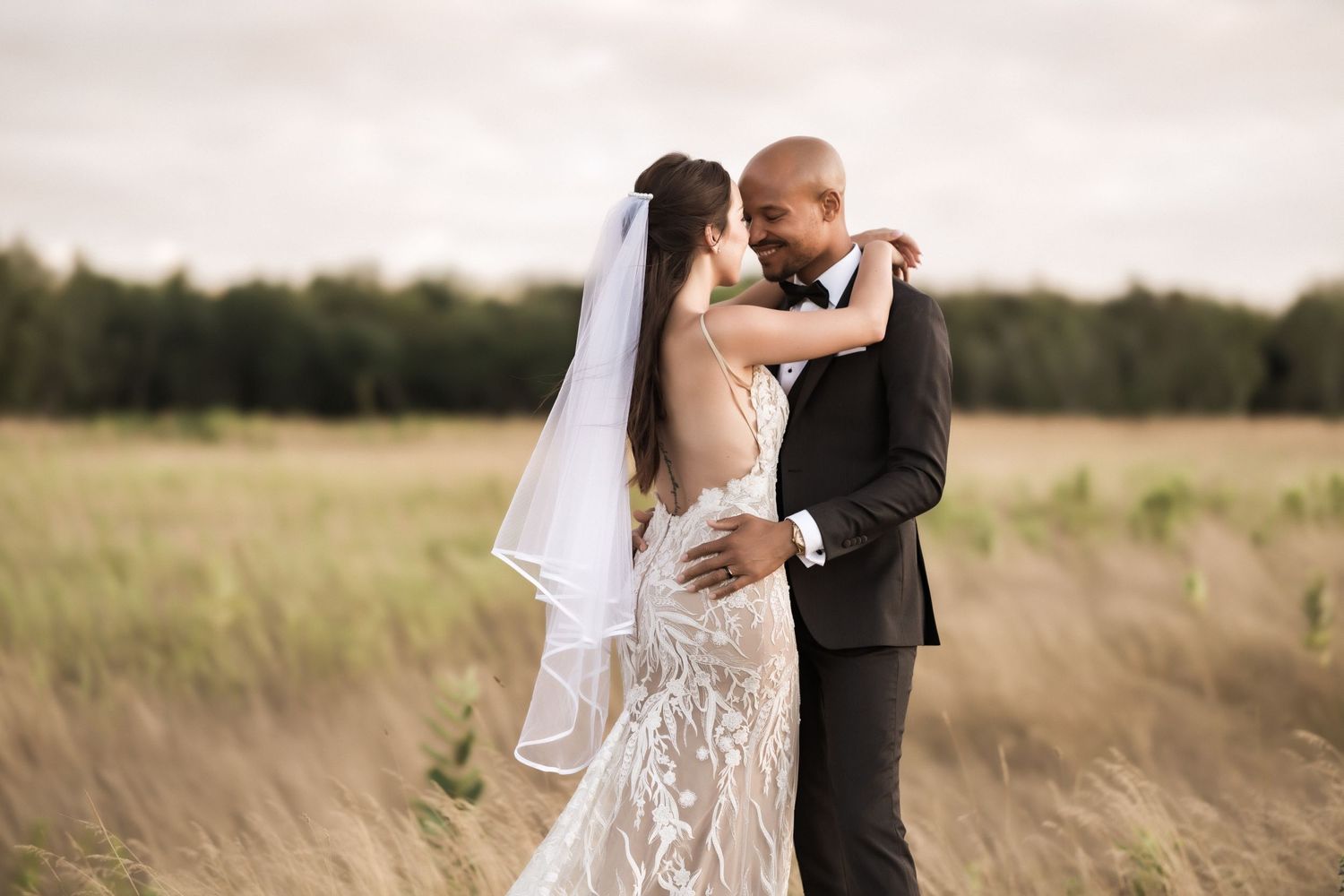 Newlyweds embrace in golden sunset light among tall grass in open field.