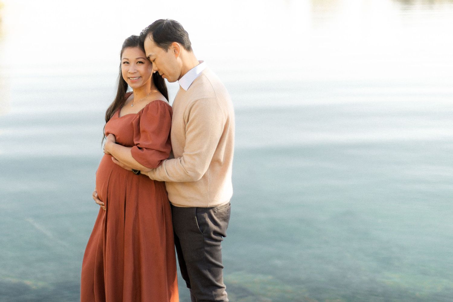 A couple shares an intimate moment against a misty coastal backdrop wearing coordinated rust and beige outfits.