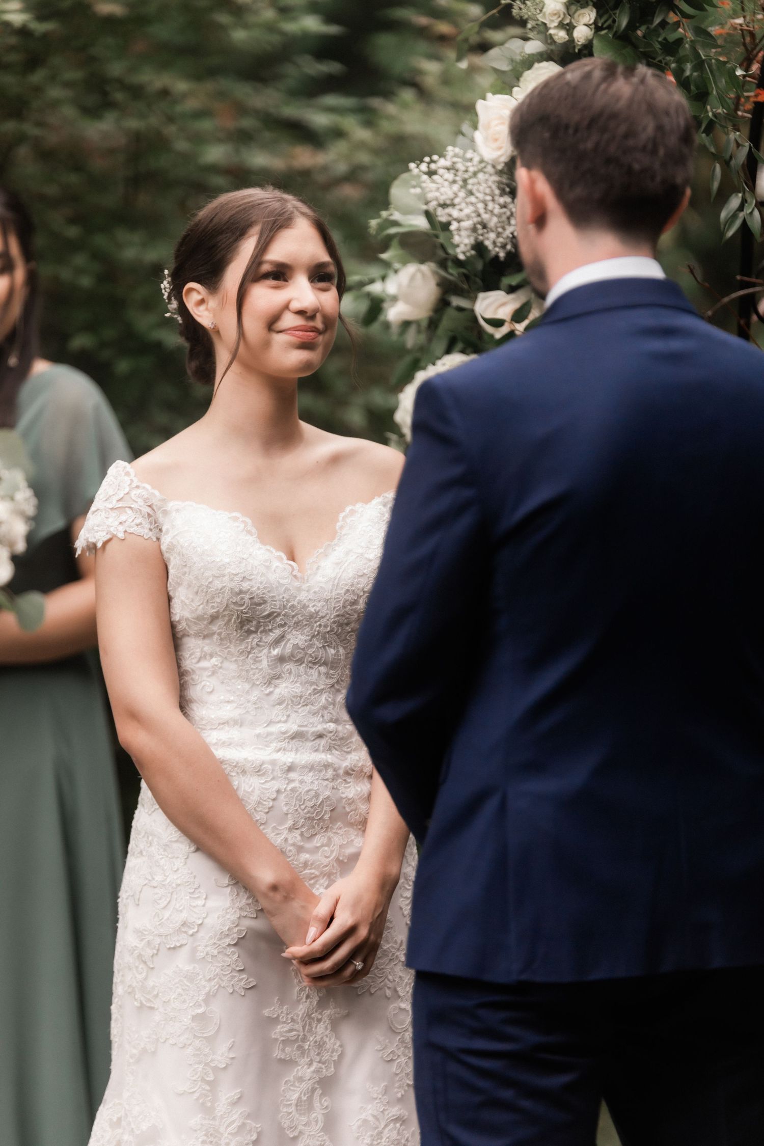 Bride in lace dress stands during outdoor garden wedding ceremony.