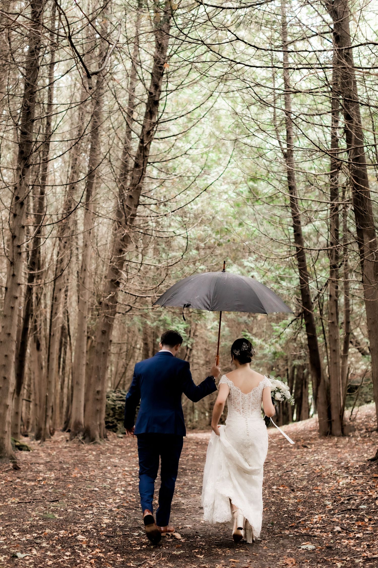 Newlyweds walking together under black umbrella on forest path covered in fallen leaves.