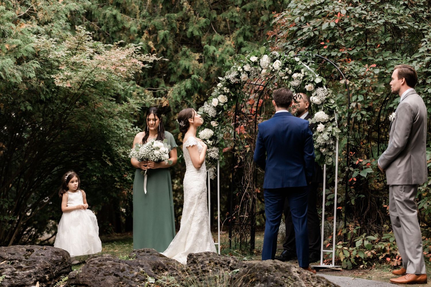 An outdoor wedding ceremony with bridesmaids in sage green dresses under a floral arch.