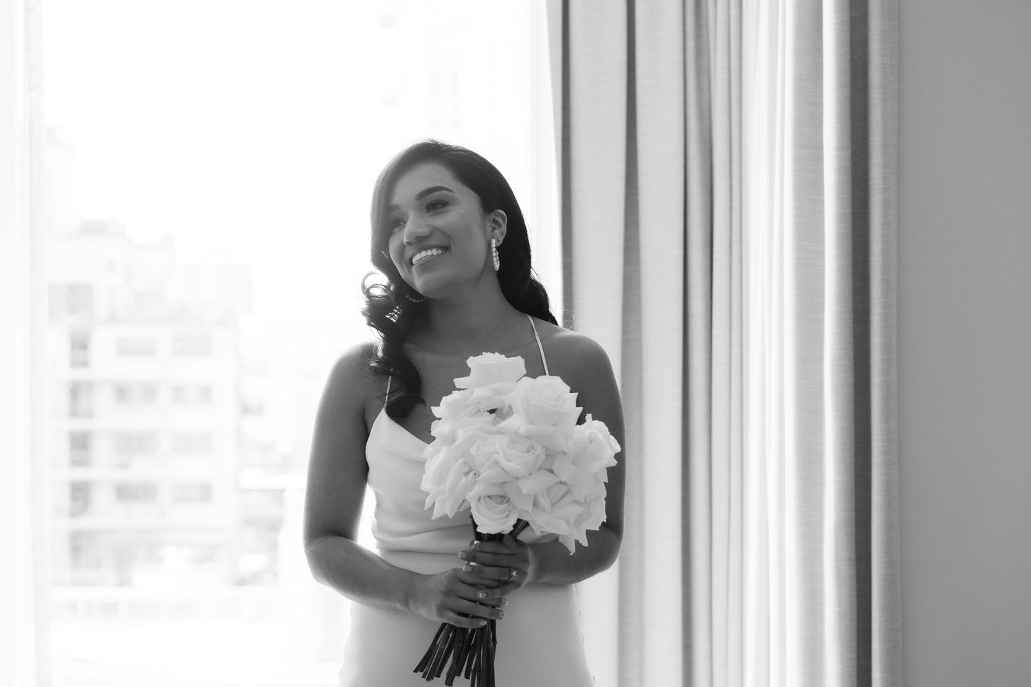 Joyful bride holding white floral bouquet by window in elegant wedding dress.